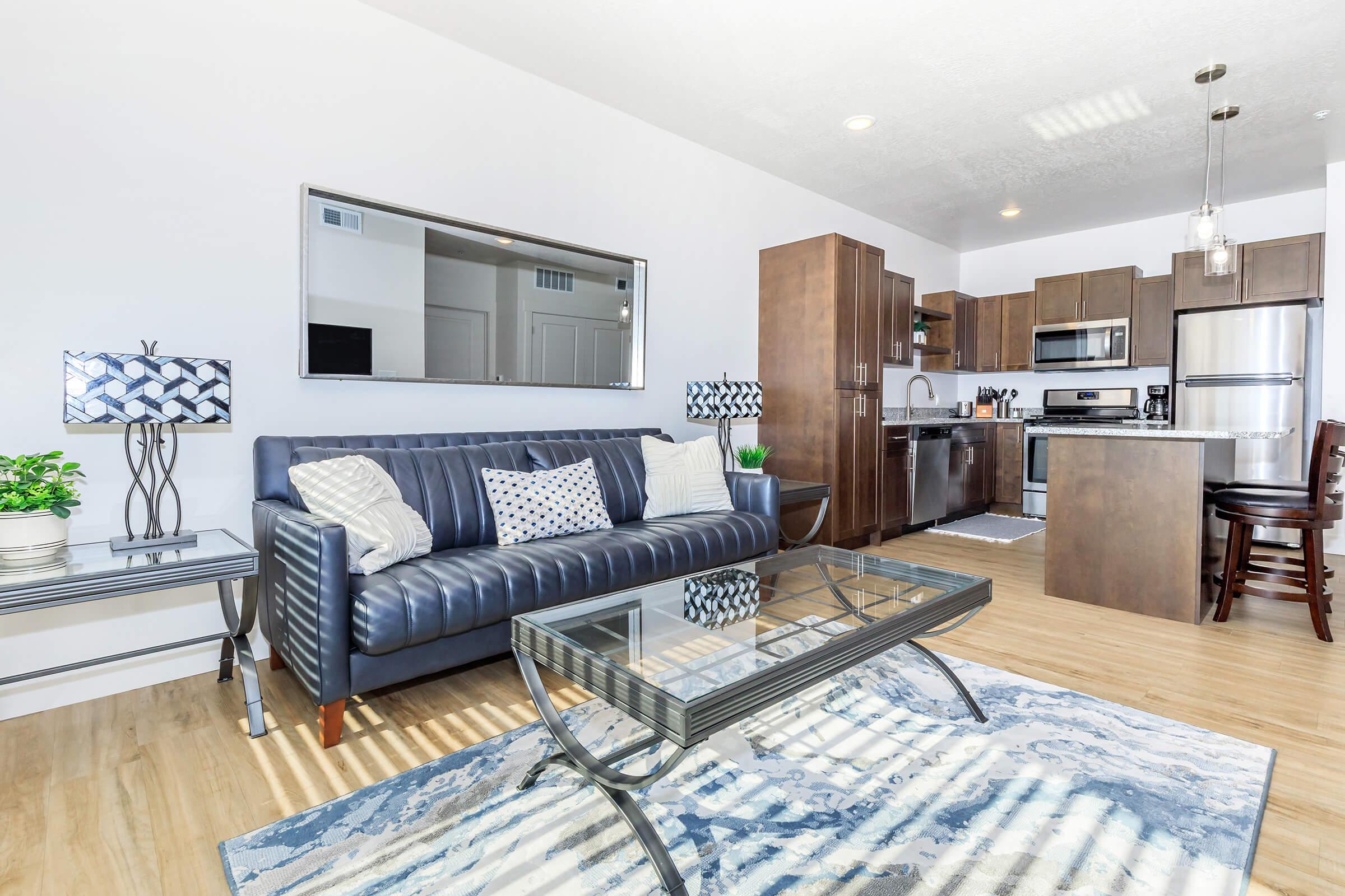 A modern living room featuring a blue leather sofa with decorative cushions, a glass coffee table on a patterned rug, and a stylish lamp on a side table. The open kitchen is visible in the background, outfitted with dark wood cabinetry and stainless steel appliances, creating a contemporary atmosphere.