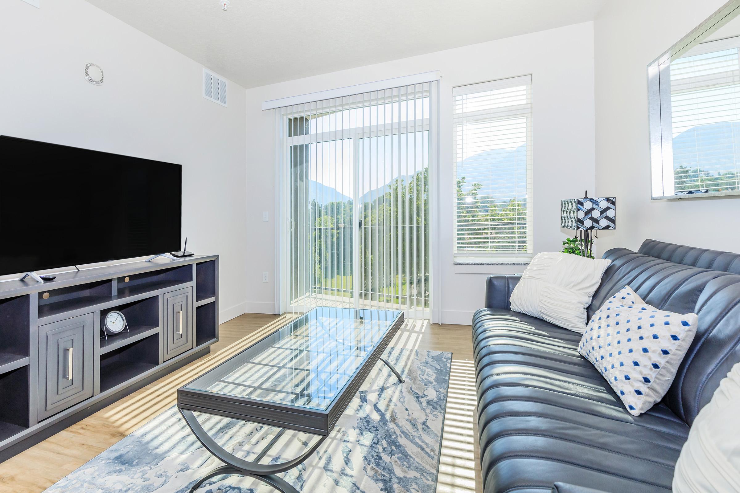 A modern living room featuring a blue sofa with white and blue decorative pillows, a glass coffee table on a patterned rug, and a large wall-mounted TV. The space is brightened by large windows with sheer blinds, offering a view of mountains and greenery outside. The décor is minimalistic and inviting.