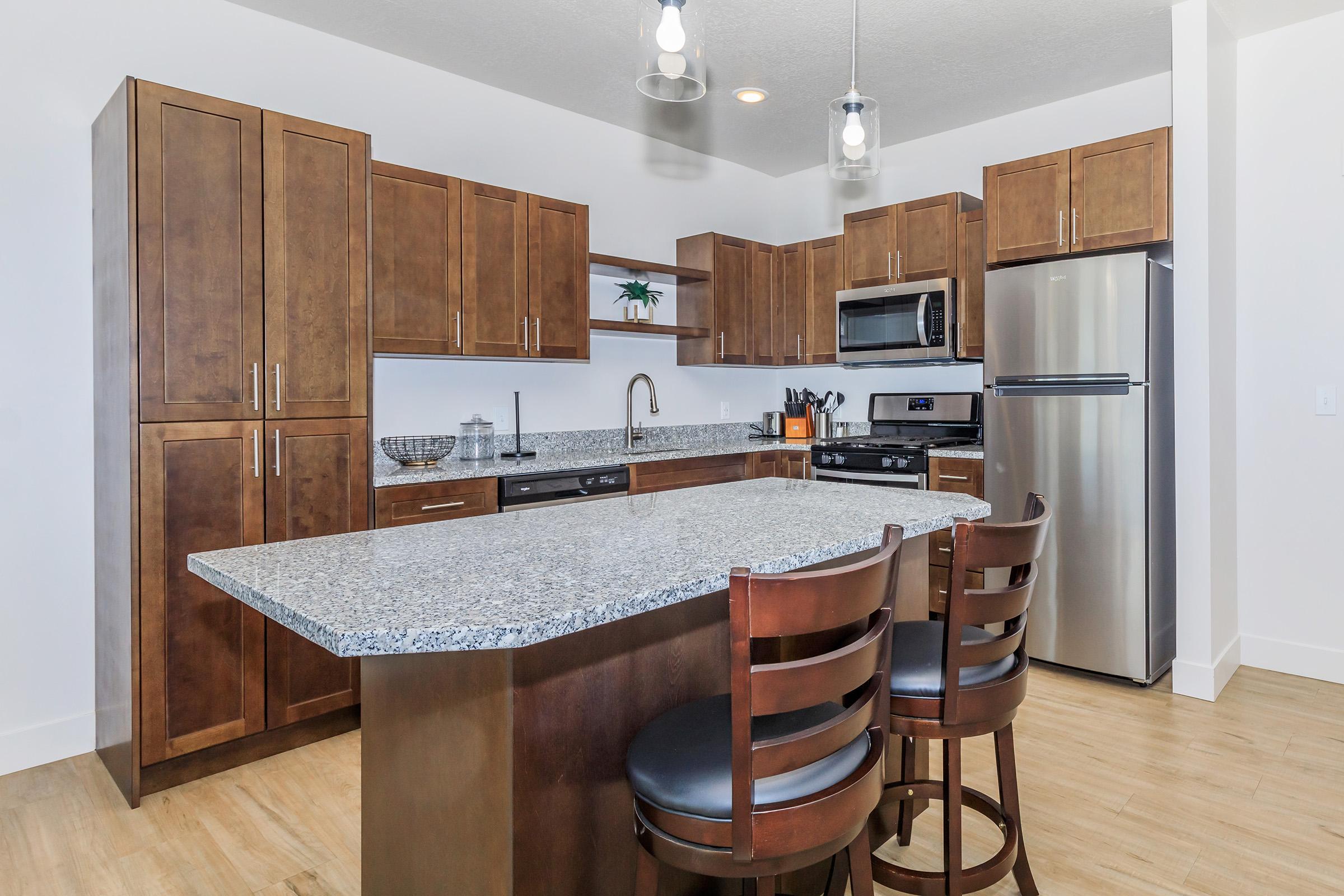 Modern kitchen featuring dark wood cabinets, a large granite island with three wooden bar stools, stainless steel appliances including a microwave and a refrigerator, and stylish pendant lighting. The space is bright, with neutral-colored walls and wood flooring, creating an inviting atmosphere.