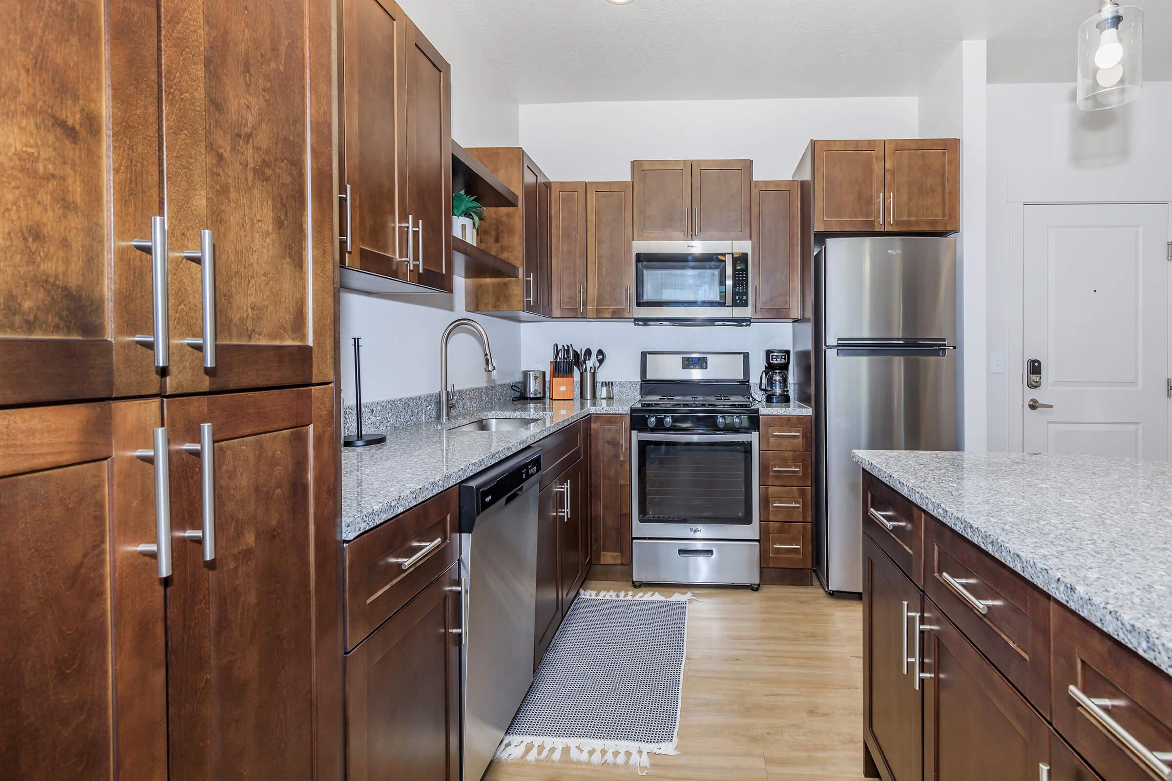 A modern kitchen featuring brown wooden cabinets, stainless steel appliances, and a granite countertop. The layout includes a dishwasher, stove, microwave, and refrigerator, with a small rug on the floor. Natural light illuminates the space, creating a warm and inviting atmosphere.