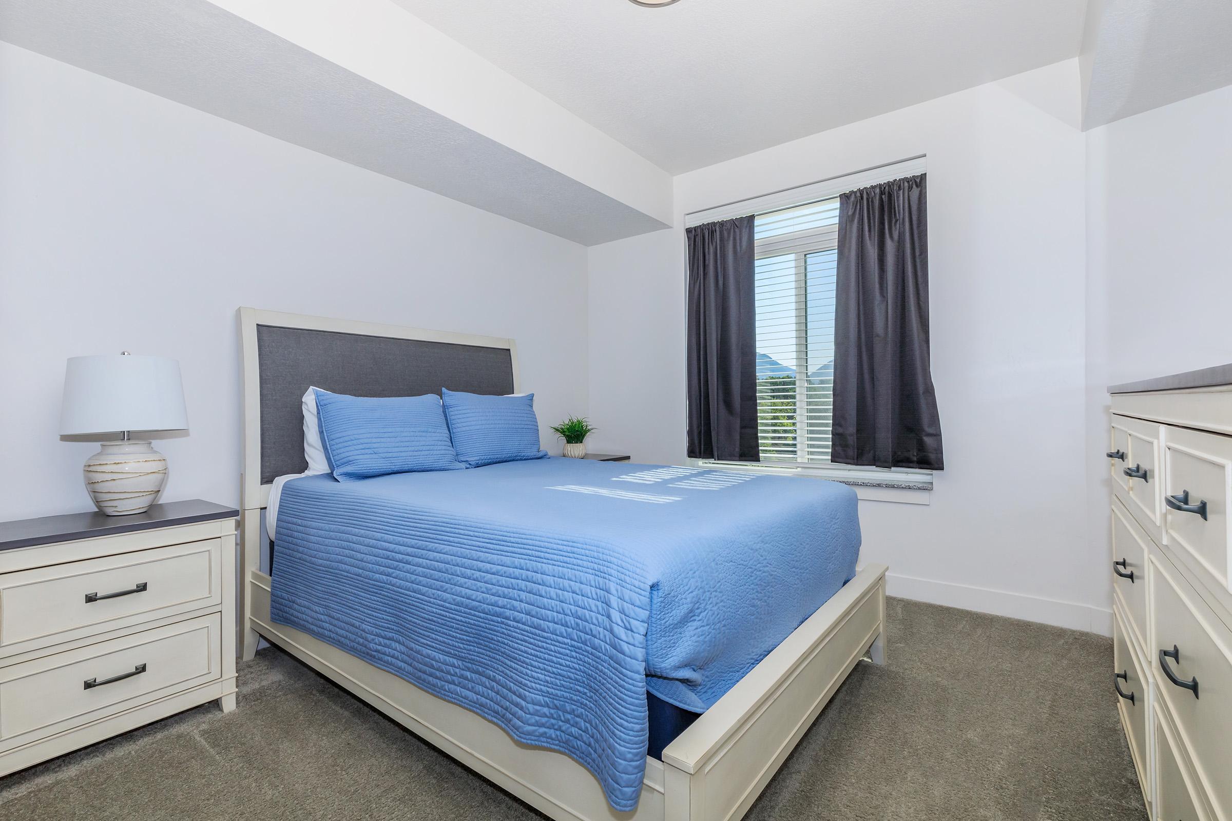 A cozy bedroom featuring a queen-sized bed with a blue quilt, light-colored nightstands with lamps, and a dresser. The room has large windows with dark curtains, allowing natural light to brighten the space. A small potted plant adds a touch of greenery to the decor.