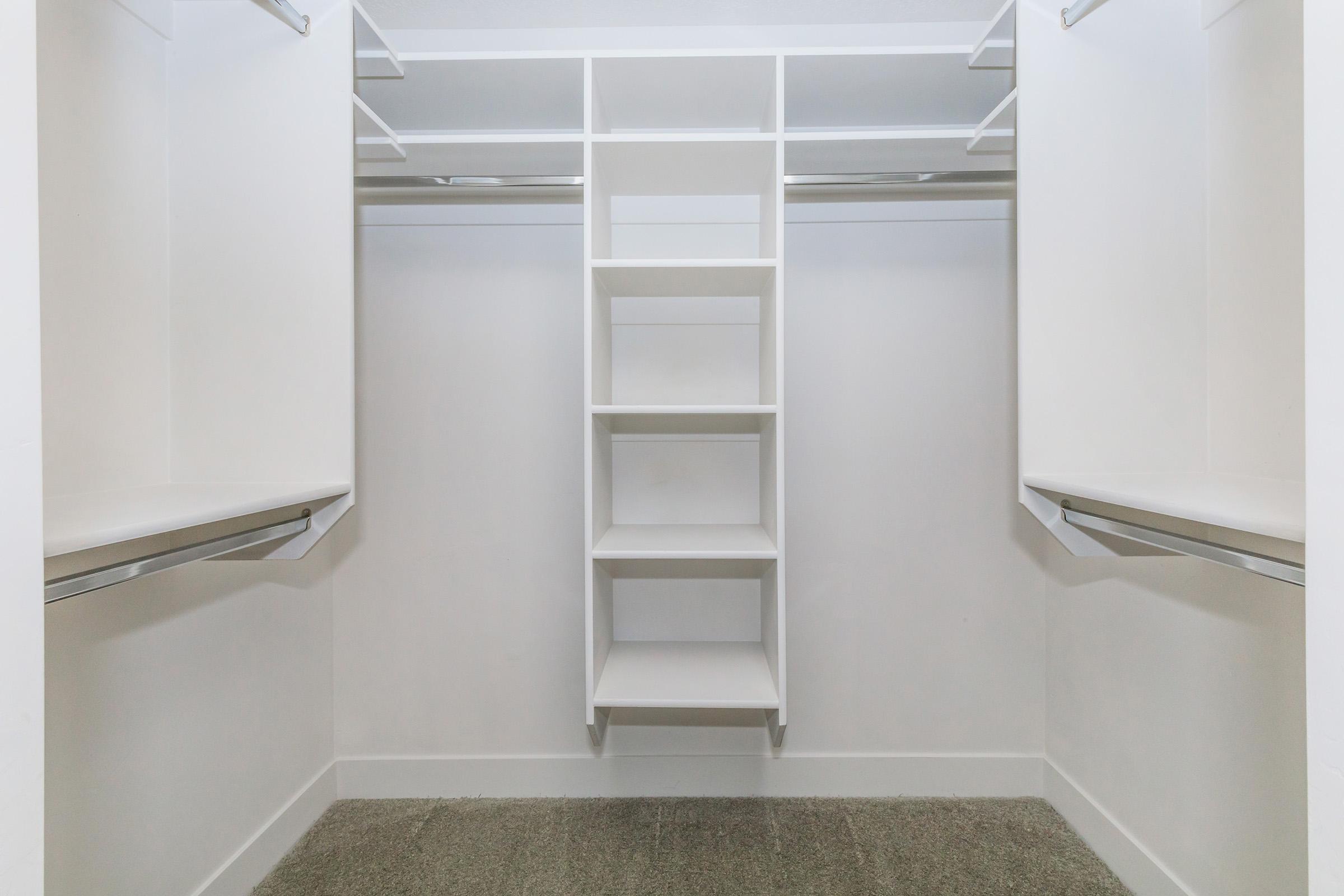 A clean, minimally styled walk-in closet featuring white shelving and hanging rods. The space is well-lit, with a carpeted floor and organized sections for clothes and storage. The design emphasizes simplicity and functionality, making it easy to access and store personal items.