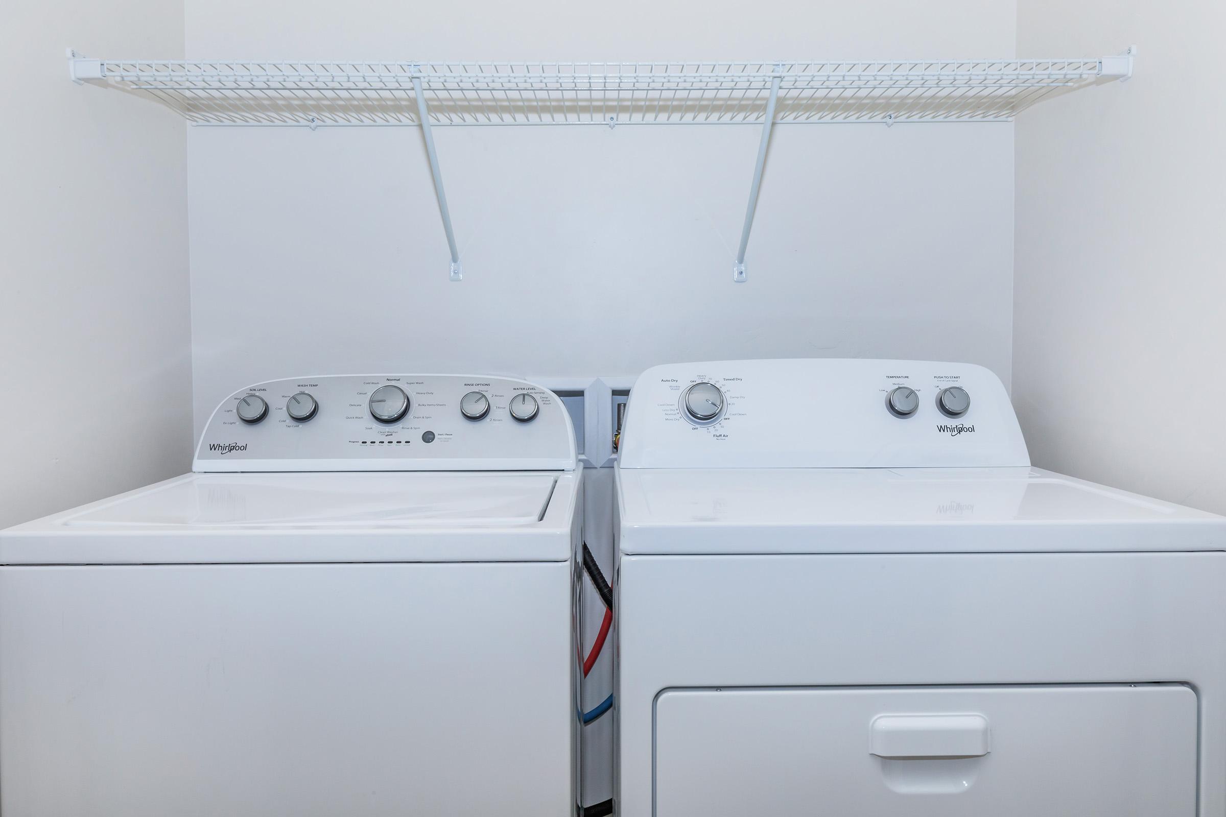 A white washer and dryer set side by side against a light-colored wall. The washing machine features control knobs and a lid, while the dryer has a similar design with its own control knobs. Above them is a white wire shelf for storage.