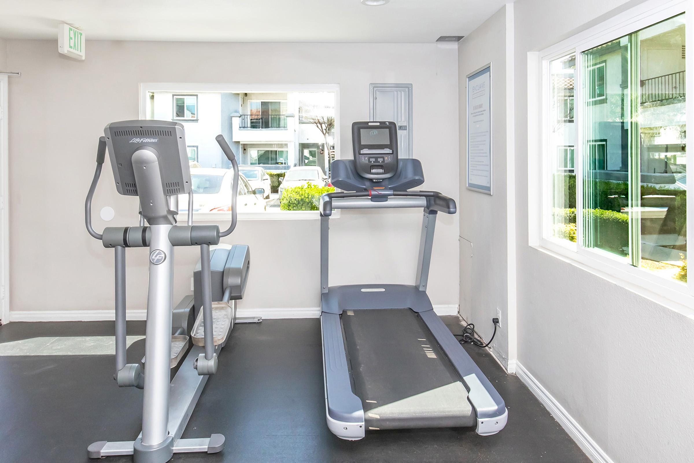 A modern gym interior featuring two pieces of exercise equipment: a stationary bike and a treadmill. The room is well-lit by natural light coming through large windows, showcasing an outdoor view. The walls are painted a light color, creating a spacious and inviting atmosphere for workouts.