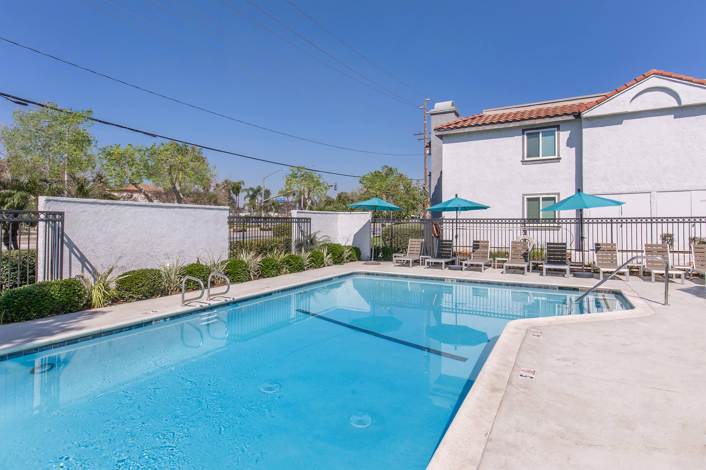 A clear, inviting swimming pool surrounded by lounge chairs and green umbrellas, with palm trees in the background. The pool area is enclosed by a fence, and a white building is visible nearby under a bright blue sky.