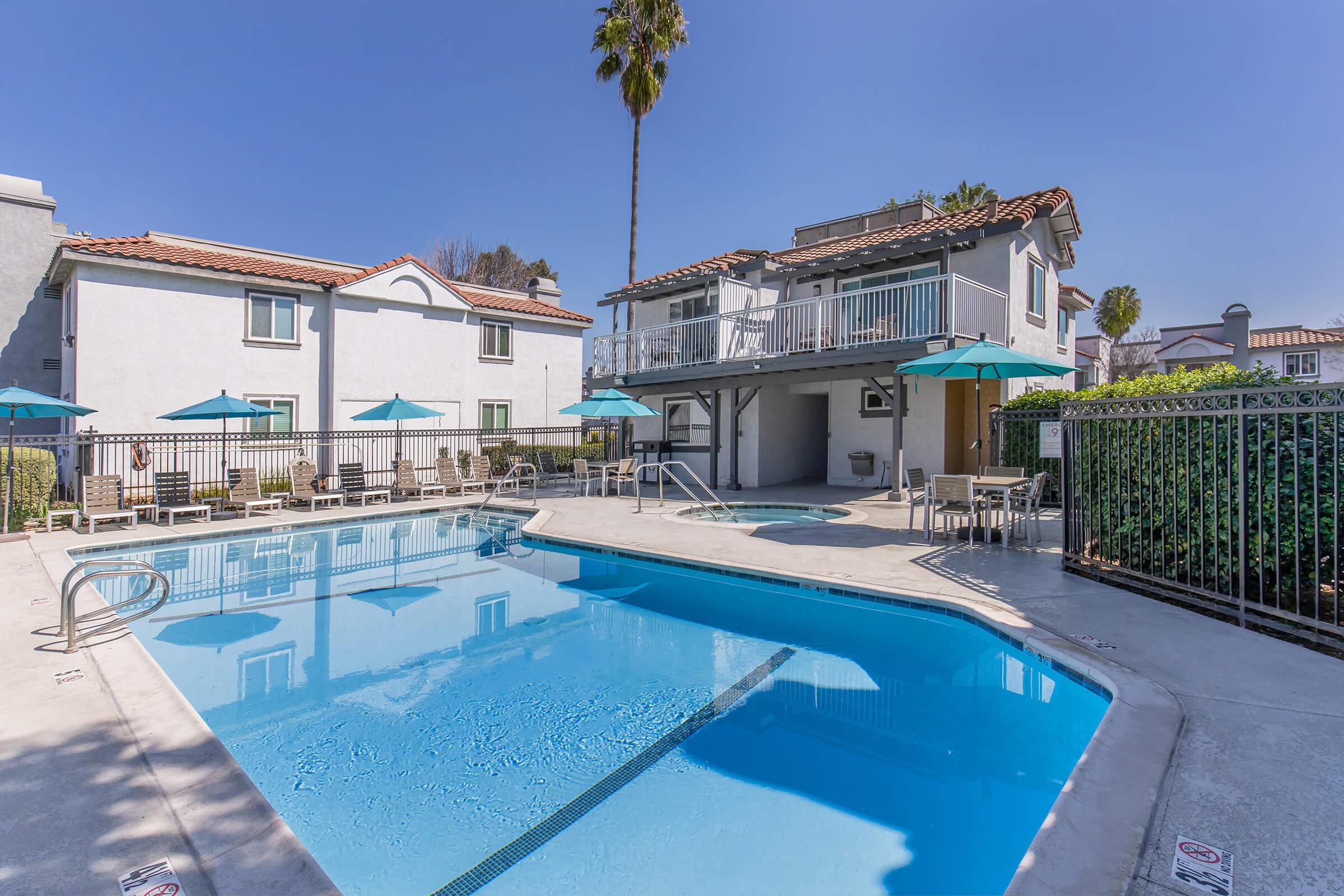 A sunny outdoor pool area featuring a clear blue swimming pool, spa, and lounge chairs. Umbrella-covered tables provide shaded seating. Surrounding the pool are well-maintained buildings with a terrace above. Palm trees and greenery enhance the relaxing atmosphere.