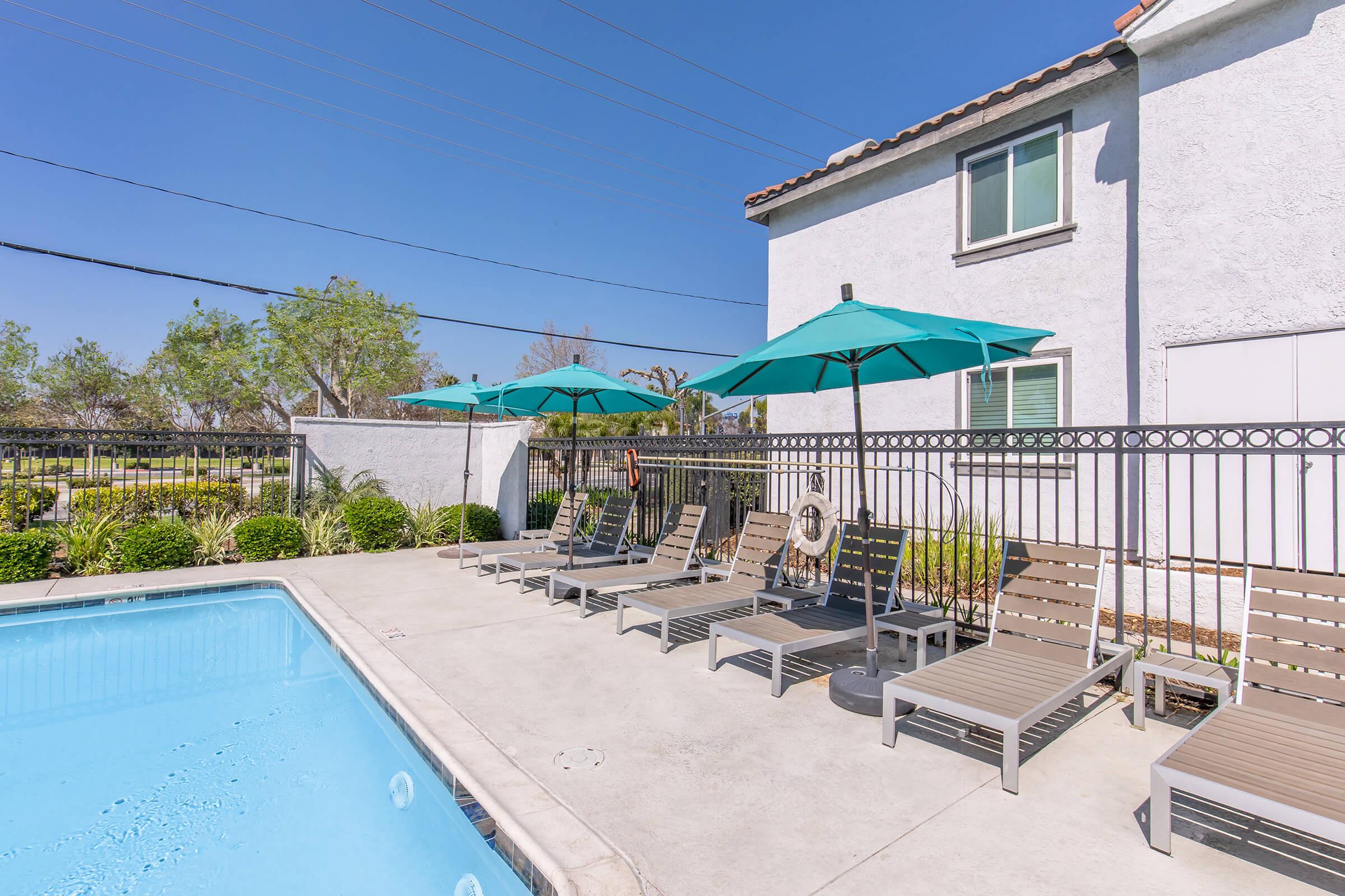 A clean swimming pool area featuring lounge chairs with teal umbrellas. The surrounding fence is visible, along with a well-maintained backyard area with trees. The building in the background is light-colored, complementing the serene atmosphere of the poolside.