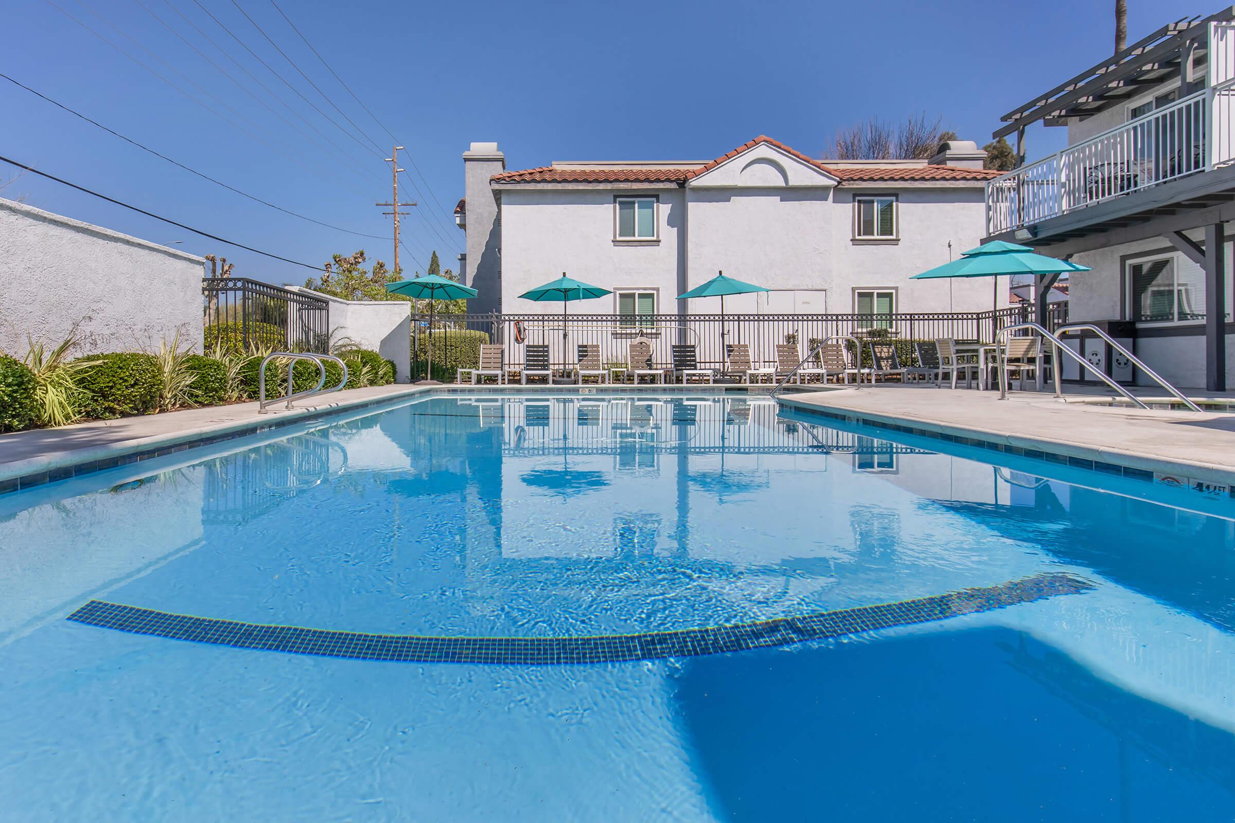 A clear swimming pool with a tiled edge, surrounded by lounge chairs and turquoise umbrellas. In the background, there is a white building with a red-tiled roof and a clear blue sky overhead. The setting appears to be a relaxing outdoor area, ideal for leisure and enjoyment.
