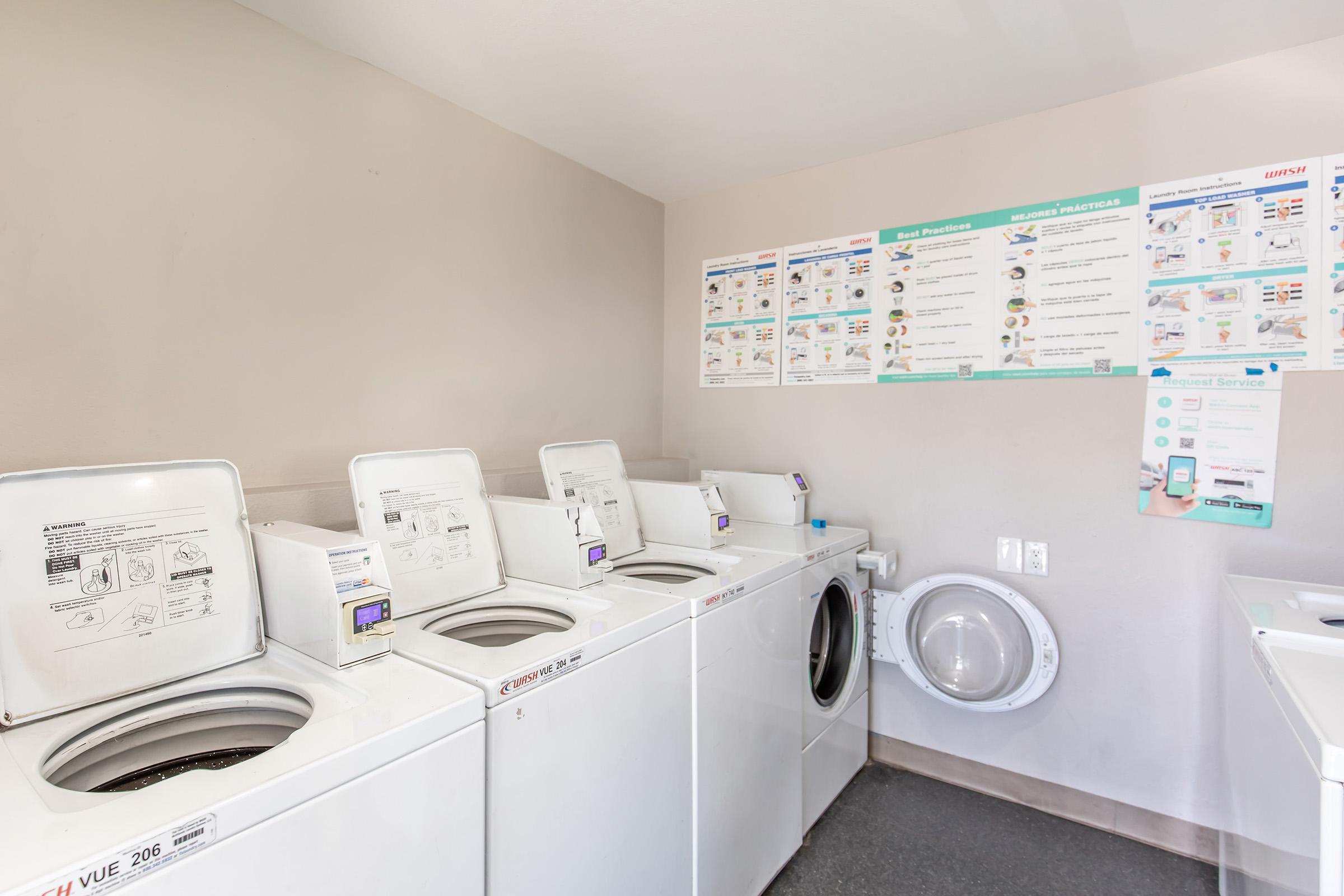 A clean laundry room featuring several washing machines and a dryer on the right. The wall is adorned with informational posters outlining laundry best practices and instructions. The space is well-lit and organized, providing a functional area for doing laundry.