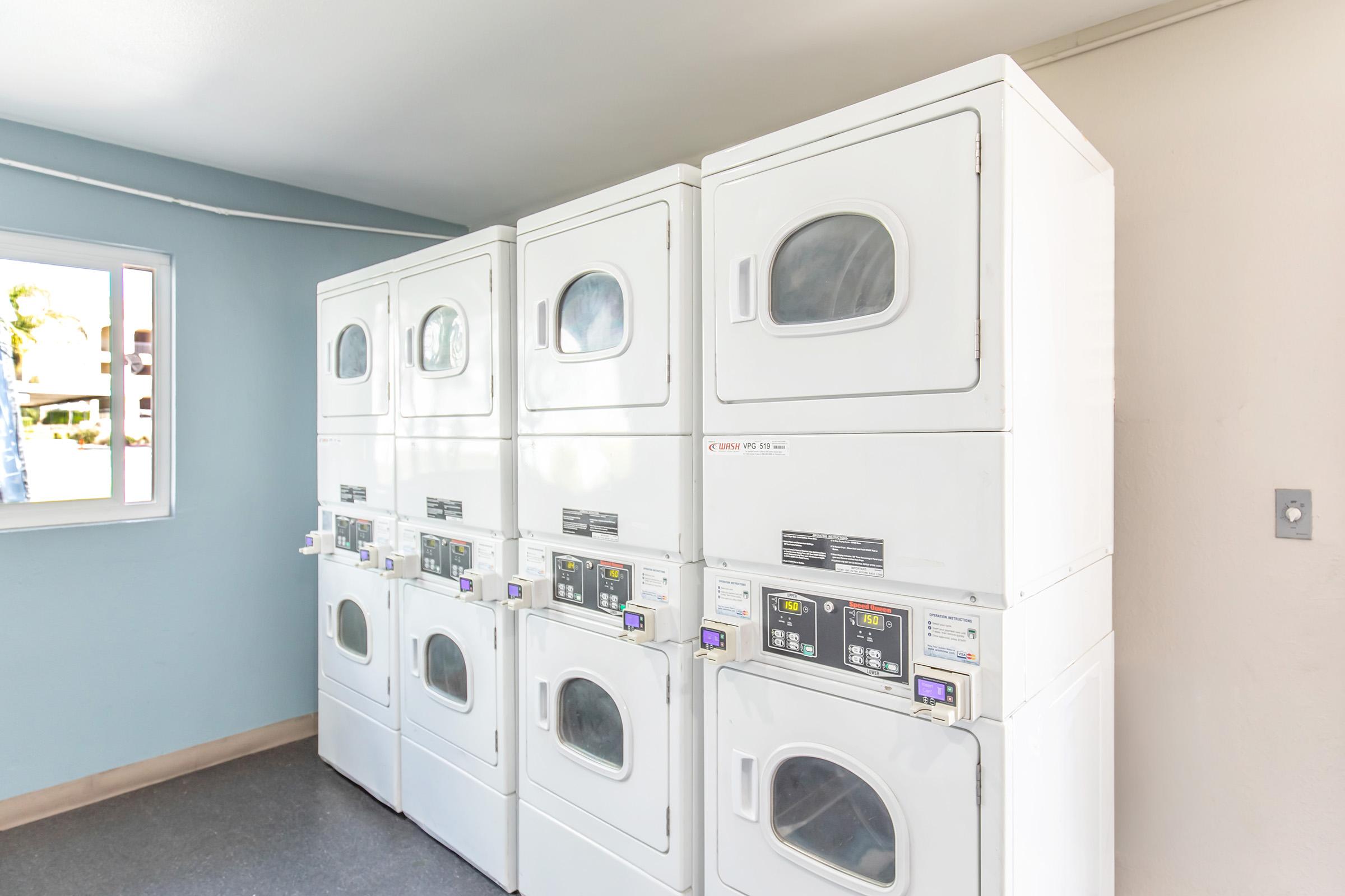 A row of six stacked commercial washing machines and dryers in a well-lit laundry room. The machines are white, and there's a window on the left side allowing natural light to enter. The walls are a light color, creating a clean and organized environment.