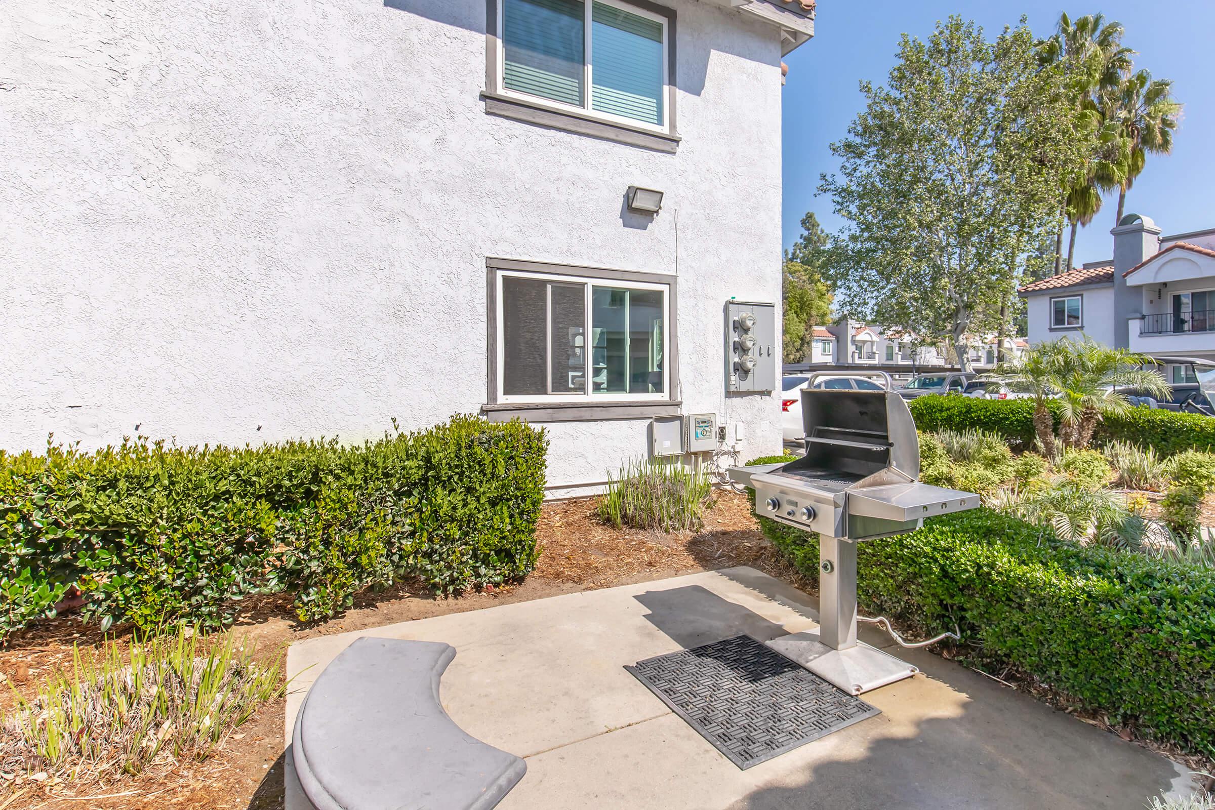 A barbecue grill on a concrete patio next to a white building, surrounded by neatly trimmed hedges and plants. There's a small bench curved around the grill area, and a clear blue sky with a few trees in the background.