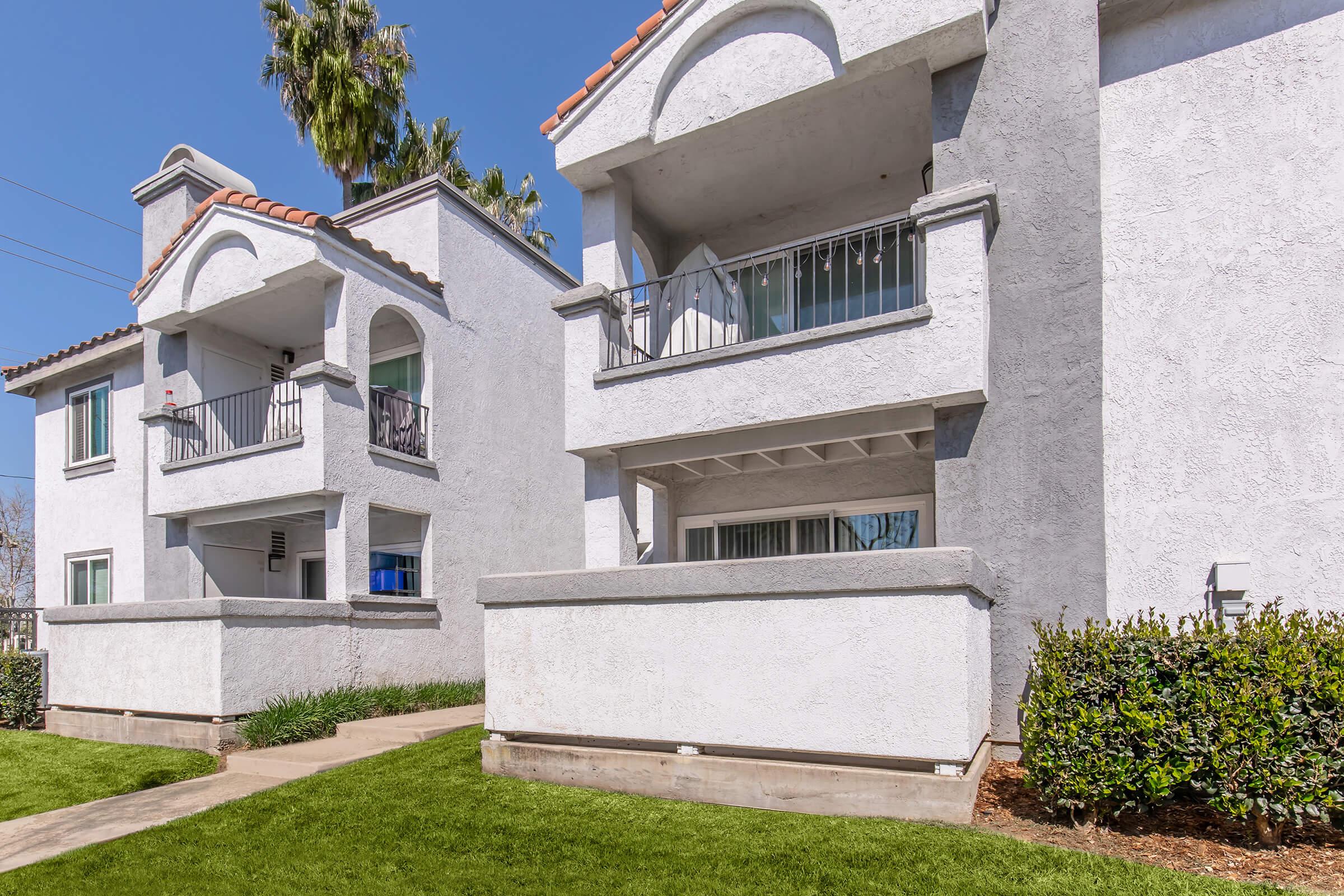 Two white, multi-family residential buildings with arched balconies. The buildings have a stucco finish and are surrounded by well-maintained green grass and shrubs. The sky is clear and blue, indicating a sunny day. Pathways lead up to the entrances of the apartments.