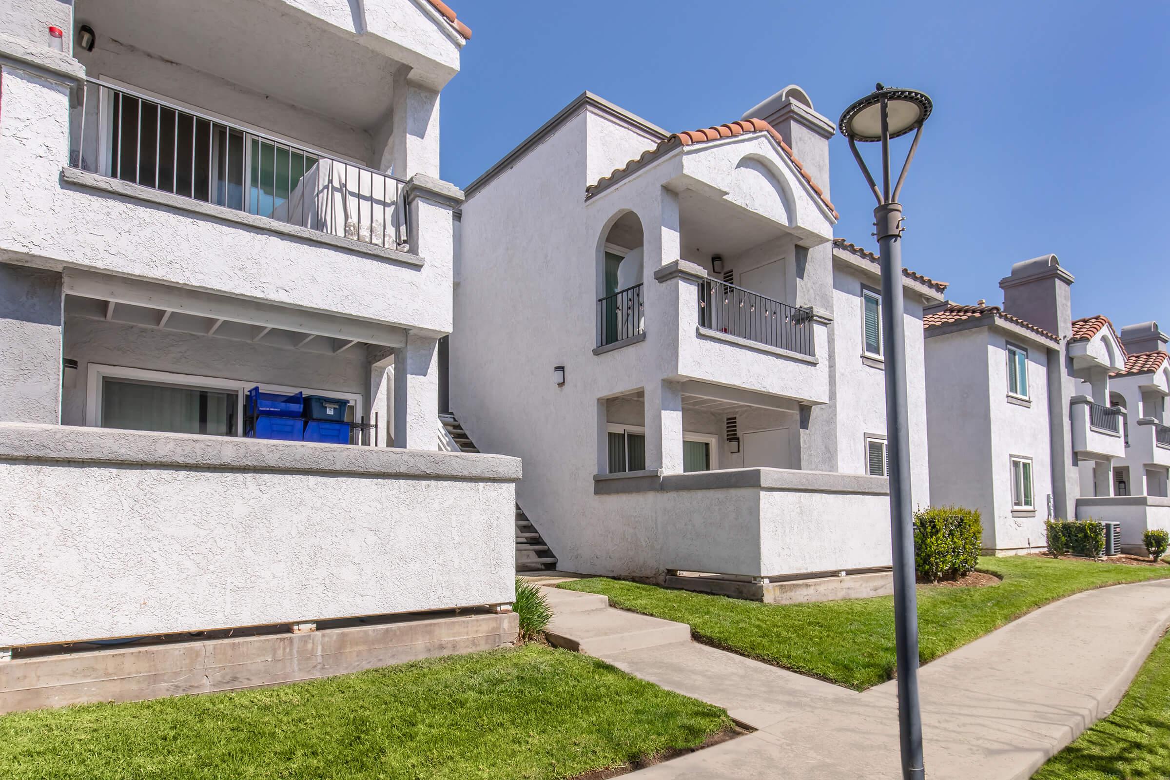 A row of two-story, white stucco apartment buildings with arched doorways and a tiled roof. The pathway in front leads to the stairs, and there is a green lawn with shrubs. A streetlamp stands along the sidewalk under a clear blue sky.