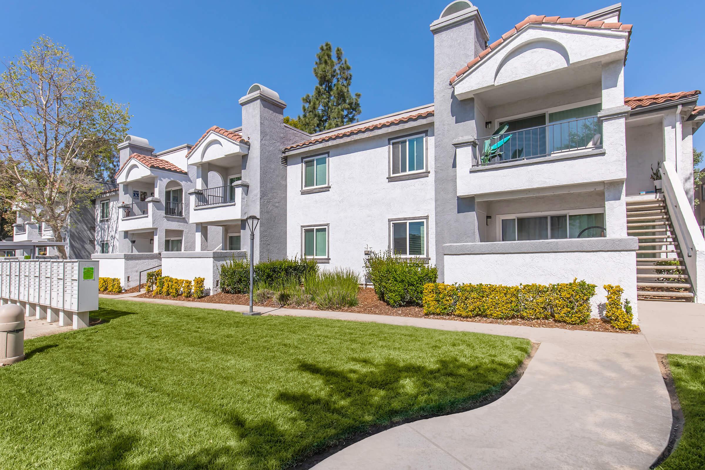 A modern two-story apartment complex with white stucco walls and terracotta roof tiles, surrounded by green lawns and landscaping. The building features balconies with sliding glass doors, and a pathway leading to the entrance. Bright blue sky and trees are visible in the background.