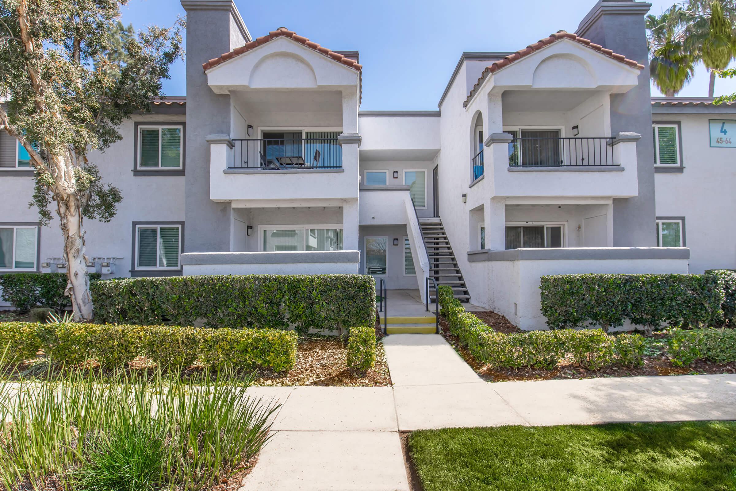 Two-story residential apartment building with a stucco exterior, featuring two balconies on the upper level, green hedges, and manicured lawns. The pathway leads to the entrance, and there are palm trees in the background under a clear blue sky.