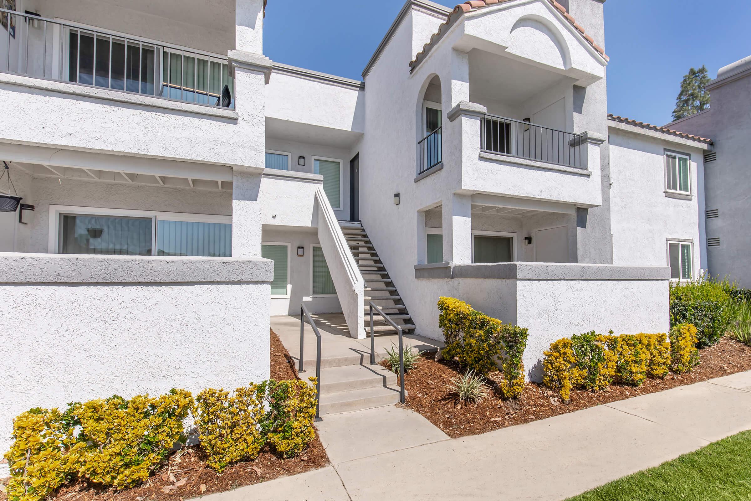 A white multi-unit residential building with two levels and a staircase leading to the second floor. The exterior features a neatly landscaped area with yellow flowering bushes and a concrete walkway. The sky is clear and blue, providing a bright atmosphere.