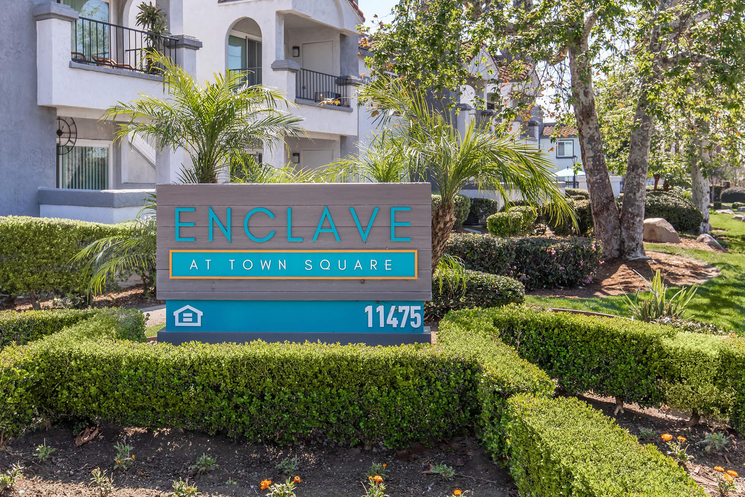 Sign for "Enclave at Town Square" with landscaped greenery and palm trees in the foreground. The sign features the address "11475" and is set against a backdrop of multi-story residential buildings.