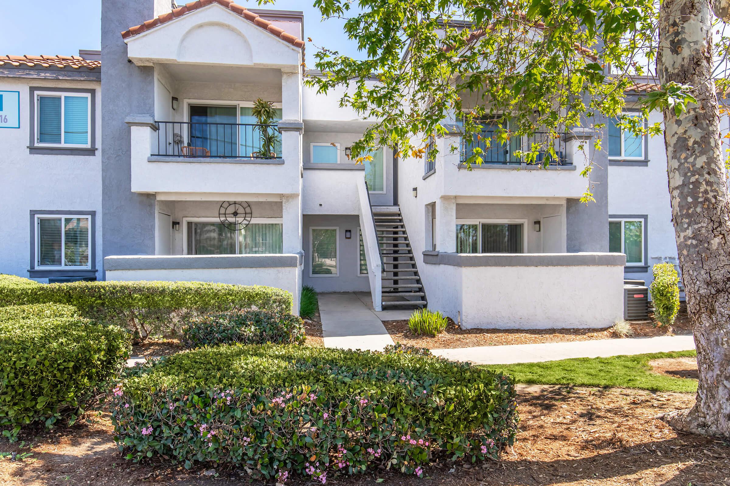 Two-story apartment building with a light gray exterior, featuring two balconies on the upper level. Lush green bushes and a tree are in the foreground, with a pathway leading up to the main entrance. The scene is sunny and well-maintained, showcasing a residential area.