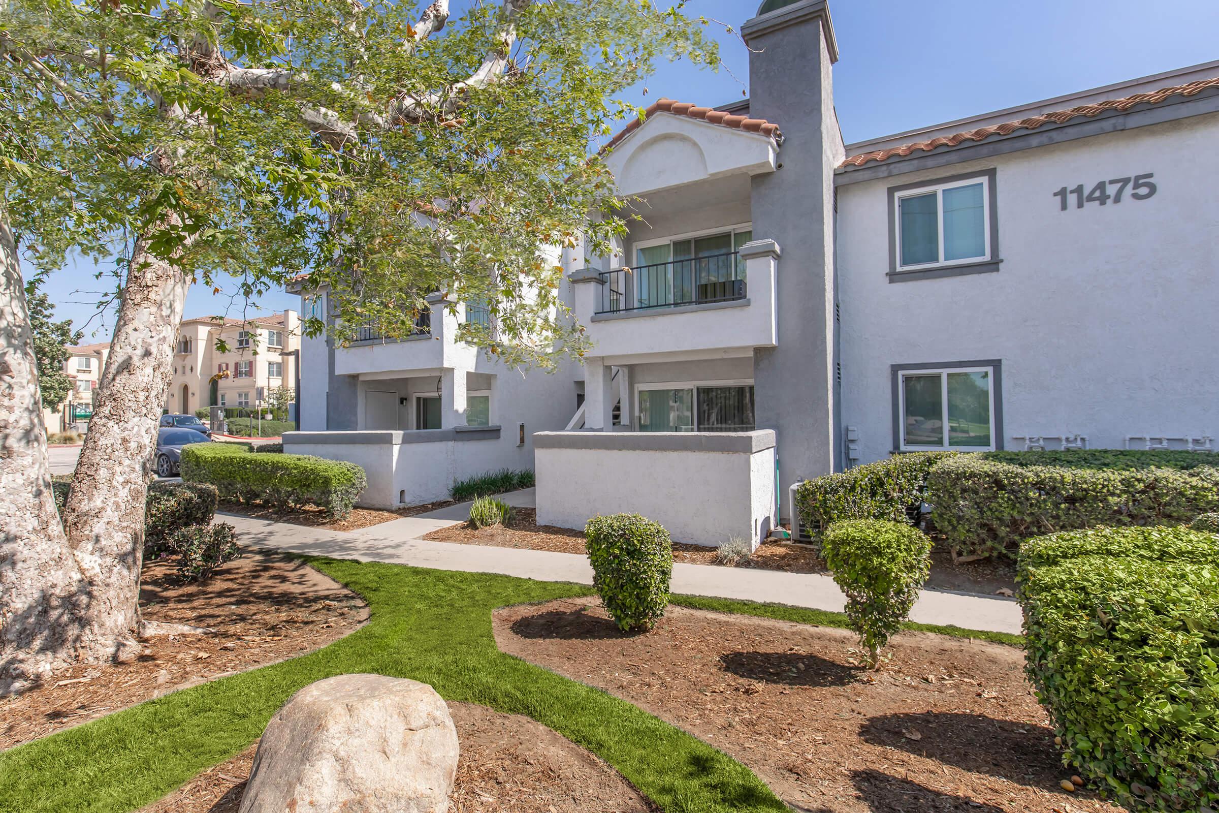 View of a residential building with a light gray exterior, featuring balconies. In the foreground, there are neatly trimmed shrubs and a large rock on a lawn. The scene is bright and sunny, showcasing a landscaped walkway leading to the entrance.