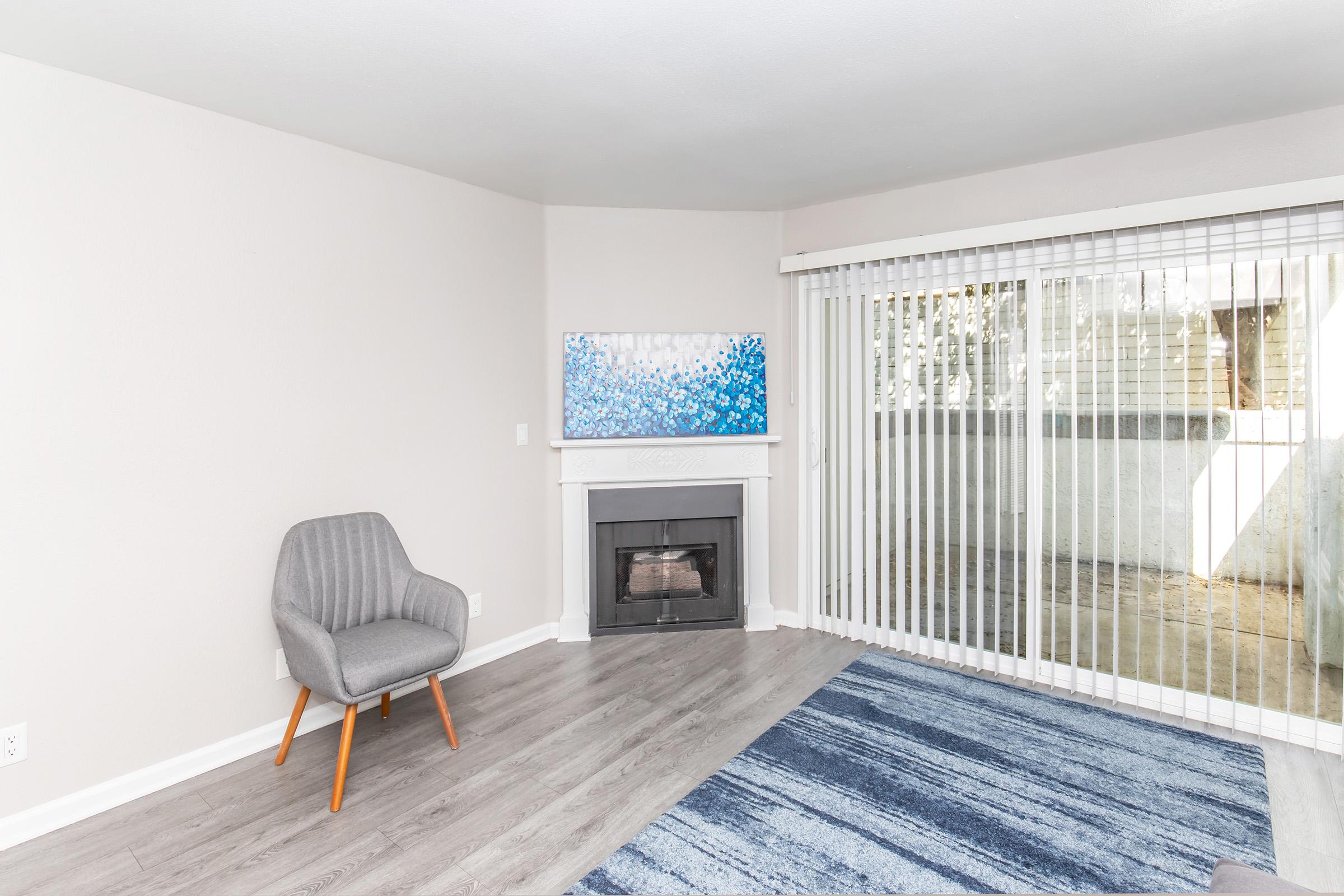 A modern living room featuring a gray armchair with wooden legs, a white fireplace, and a large wall-mounted television displaying abstract blue artwork. The room has light-colored walls and a sliding glass door covered by vertical blinds, allowing natural light to brighten the space. A blue area rug is placed on the floor.