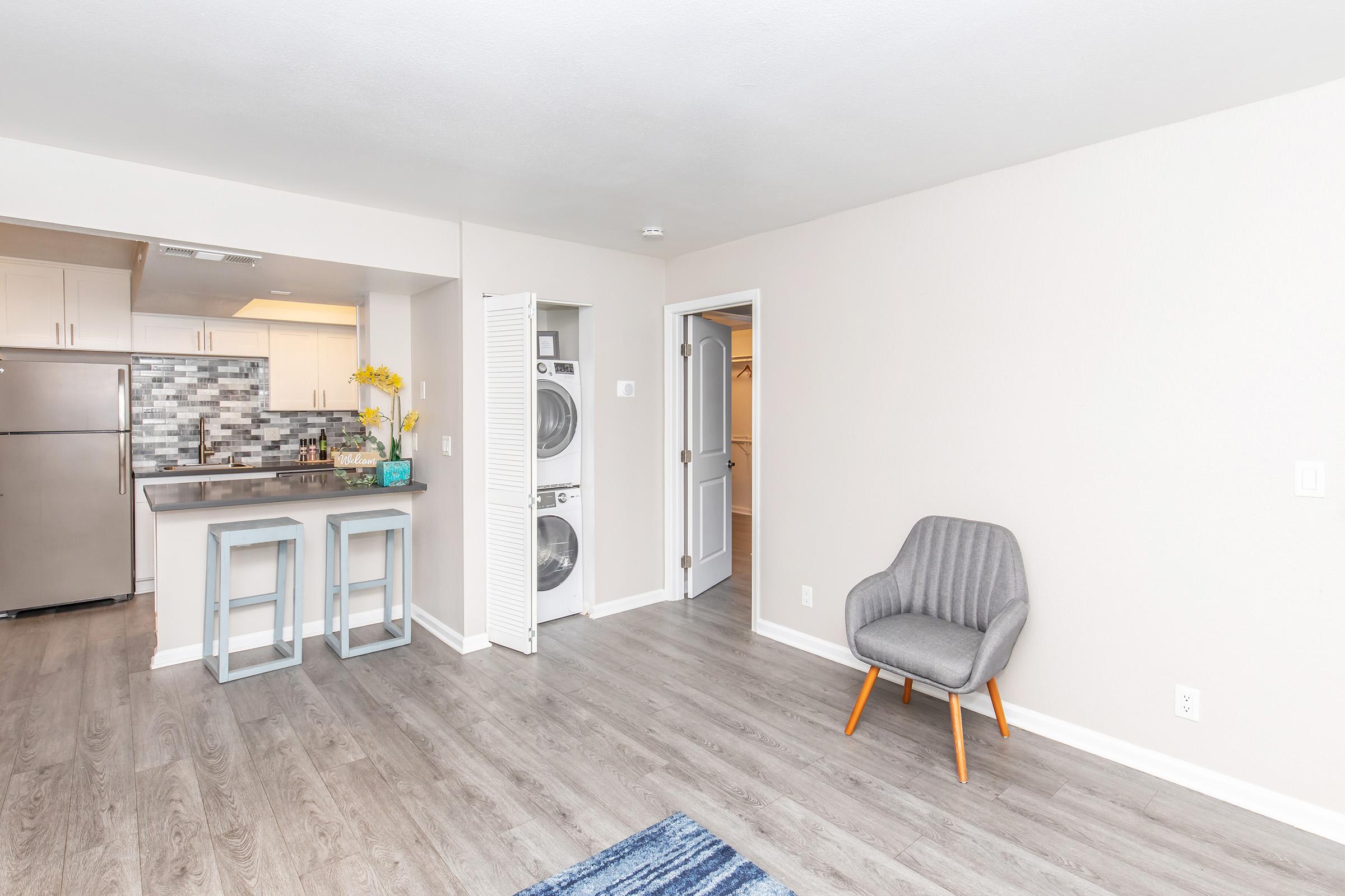 A spacious, modern apartment interior featuring a kitchen area with stainless steel appliances and a tiled backsplash. There’s a small breakfast bar with two stools, a cozy gray chair, and a laundry closet with stacked washer and dryer. Light walls and laminate flooring create a bright atmosphere.