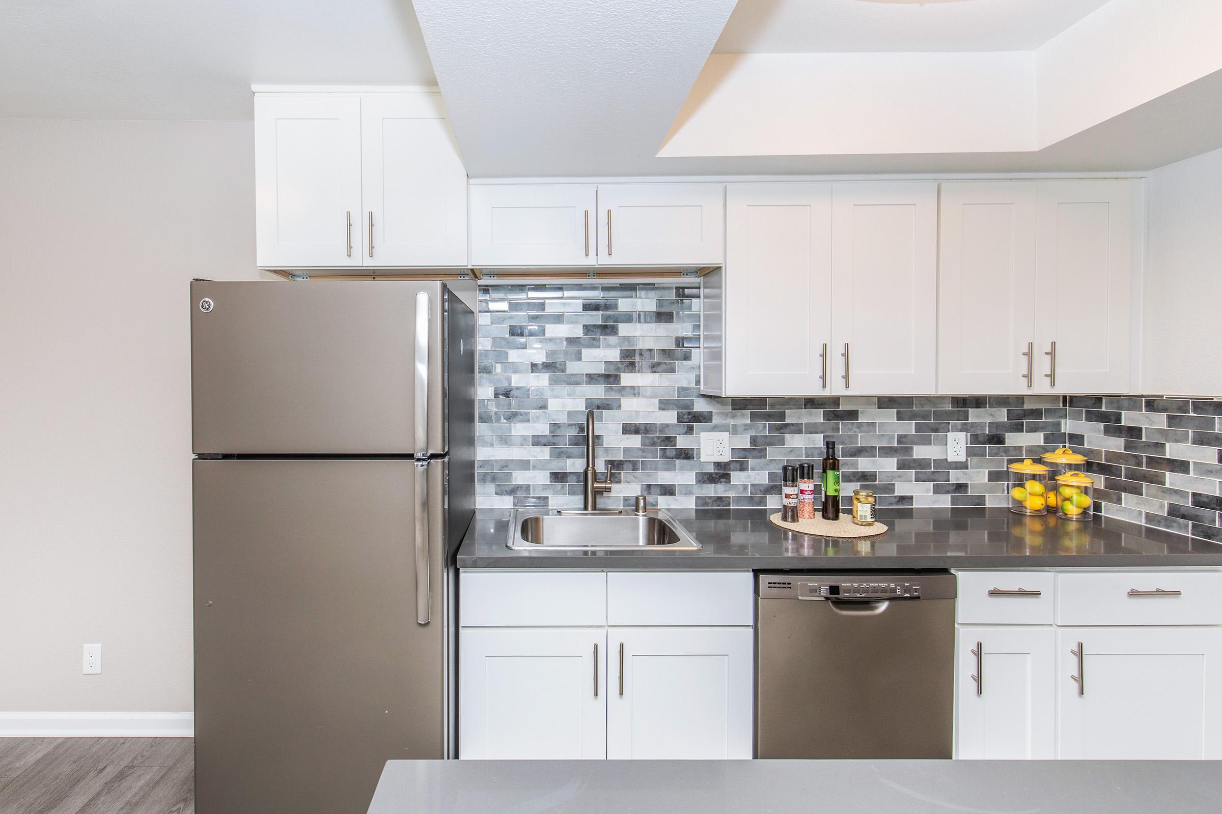 Modern kitchen featuring stainless steel appliances, including a fridge and dishwasher. The design includes white cabinets and a sleek countertop with a gray and black tile backsplash. A bowl of lemons is displayed on the counter, adding a touch of color to the contemporary space.