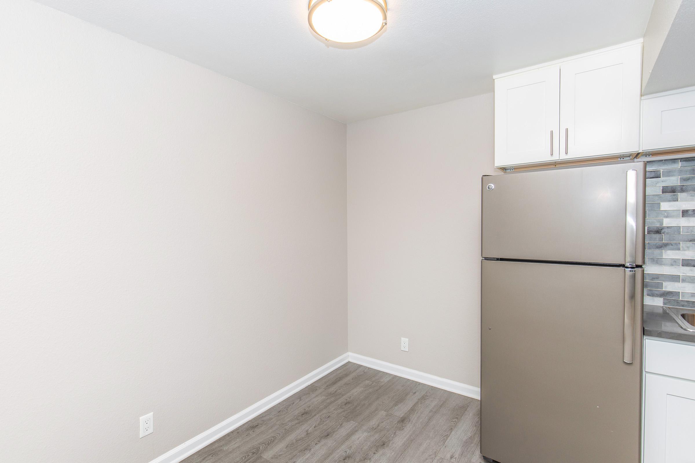 A clean, modern kitchen corner featuring a stainless steel refrigerator, light gray walls, and newly installed flooring. The area is well-lit by a ceiling fixture, and there's cabinetry above the refrigerator. The space has a minimalist design, emphasizing functionality and style.