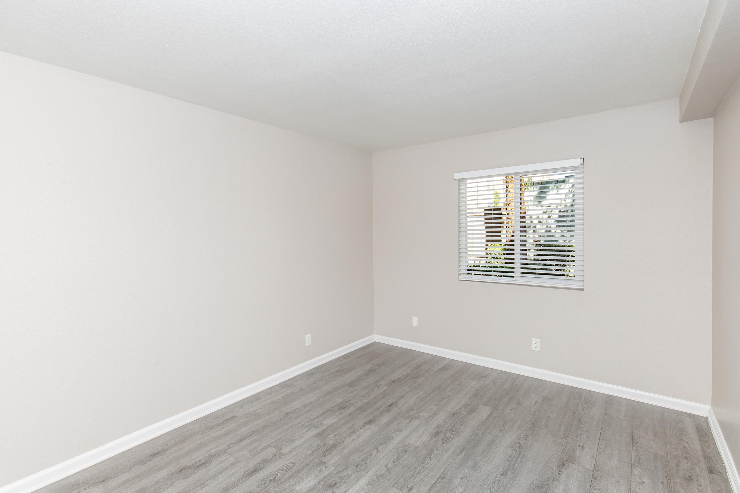 A vacant room with light-colored walls and a wood-style floor. There is a window with blinds, allowing natural light to enter. The walls are bare, creating a minimalistic and spacious atmosphere.