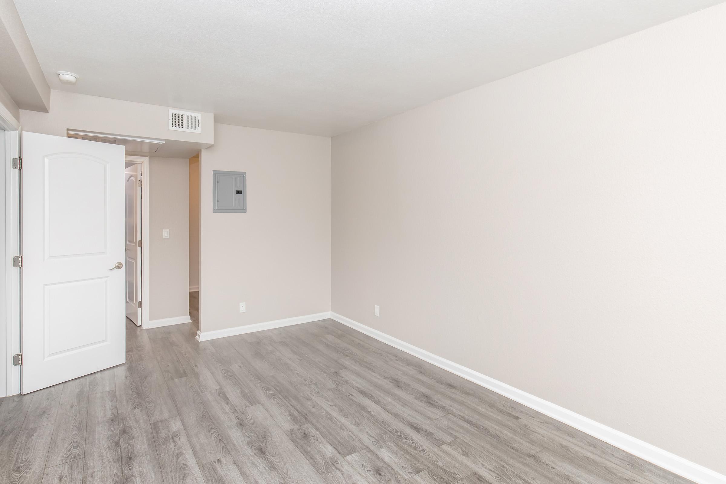 Empty room with light-colored walls and a wooden floor. A white door is partially visible on the left, leading to another space. There is an electrical panel on the wall and a small closet entrance visible in the background. The overall atmosphere is clean and modern.
