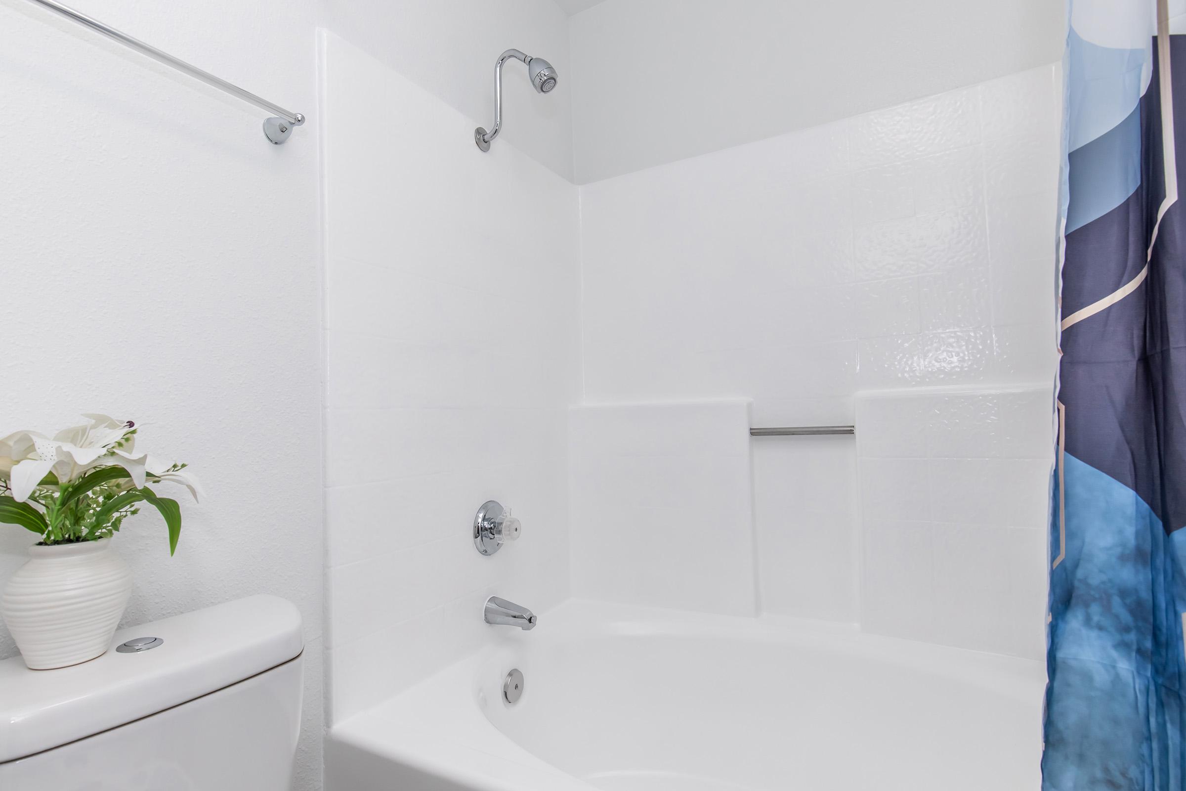 A clean and modern bathroom featuring a white bathtub with a chrome faucet, a toilet with a matching tank, and a shower area with a blue and white patterned curtain. A small vase with flowers adds a touch of decor to the space. The walls are light-colored, enhancing the brightness of the room.