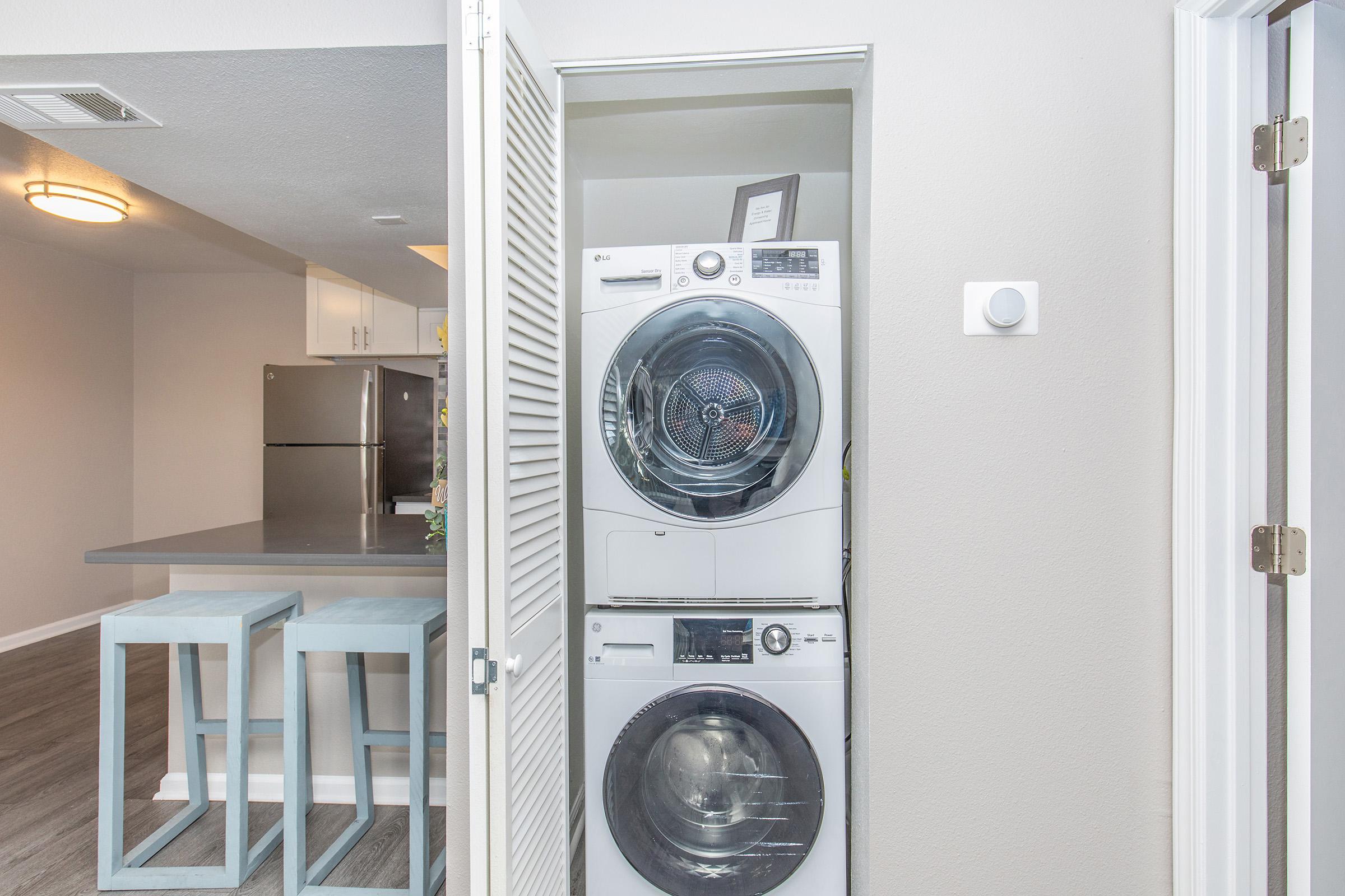 A laundry area featuring a stacked washer and dryer units behind a white louvered door. In the background, a modern kitchen with a gray countertop, bar stools, and stainless steel appliances is visible. The walls are painted in a light color and the flooring is a wood-like laminate.