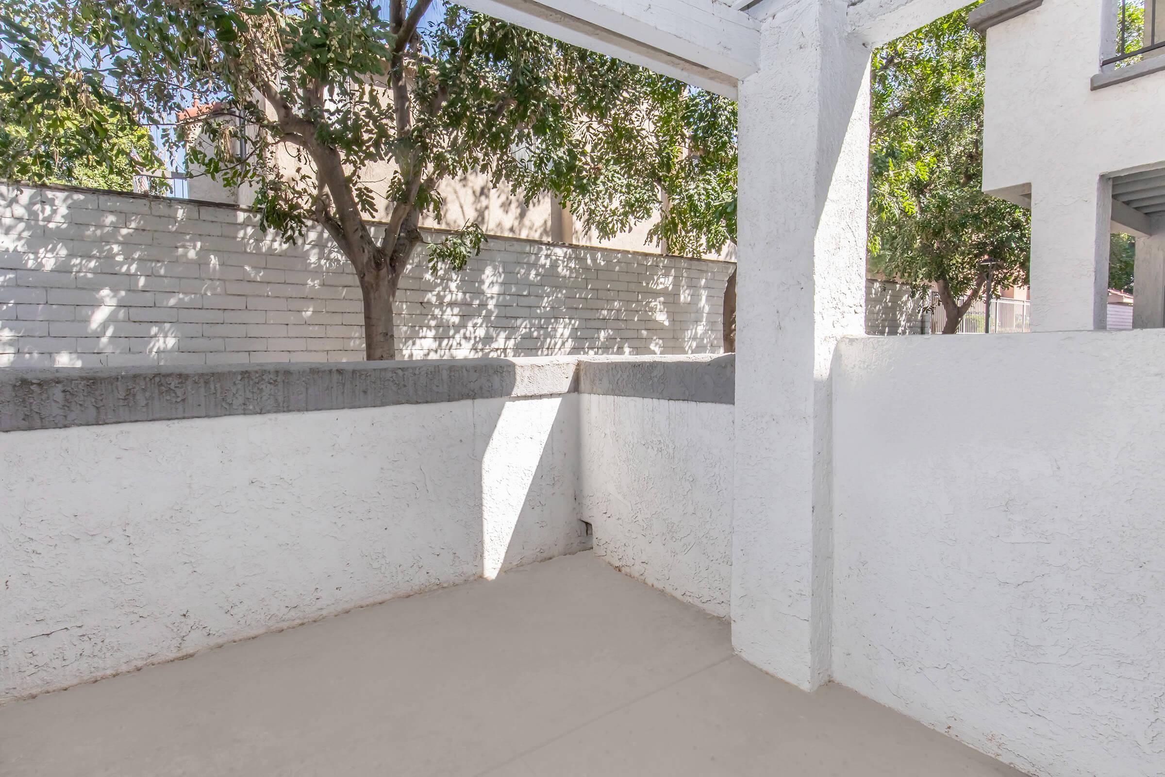 A vacant outdoor space with white walls and a concrete floor, partially shaded by trees. Sunlight creates a diagonal shadow on the ground, and there is a clear blue sky visible above. The area is enclosed, offering a private, serene atmosphere.