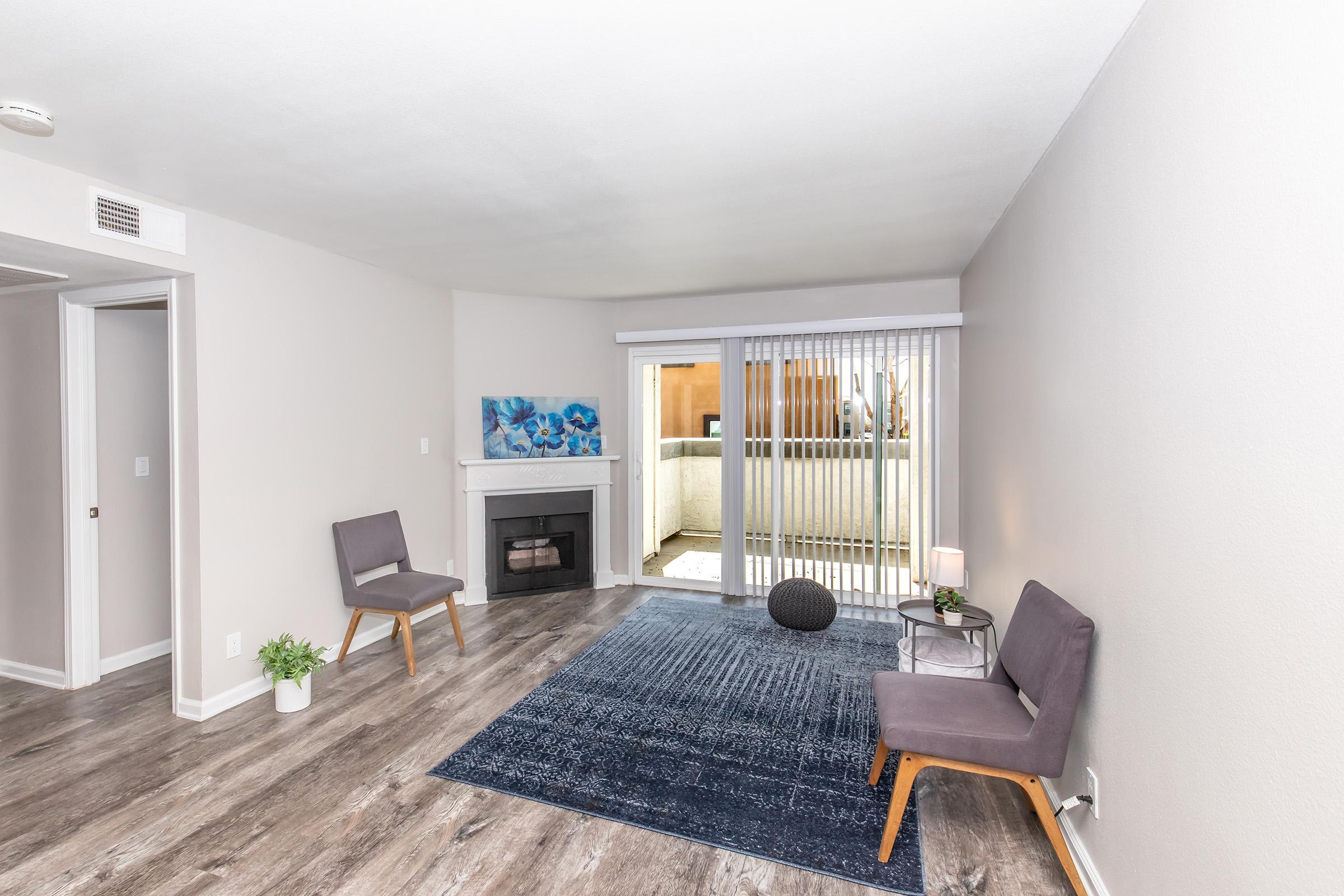 A cozy living room featuring two gray chairs, a dark blue rug, a small potted plant, and a fireplace. Large sliding glass doors lead to a balcony with outdoor seating. The walls are painted in a light tone, creating a bright and inviting atmosphere.