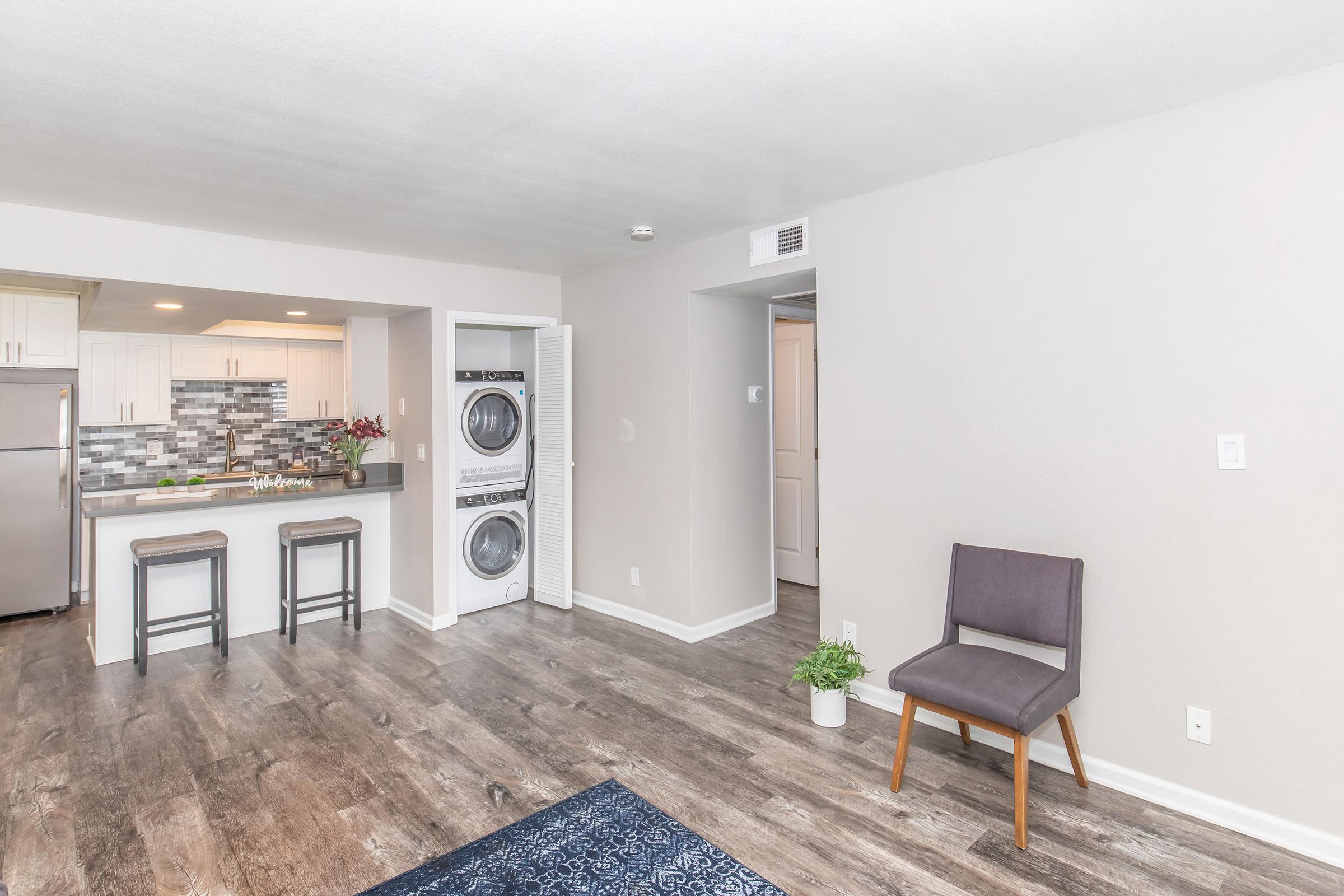 A modern, open-concept living space featuring a kitchen with a gray and white tile backsplash, stainless steel appliances, and wooden counter stools. To the side, a laundry area with stacked washer and dryer, complemented by a small potted plant and a single brown chair on a wooden floor.