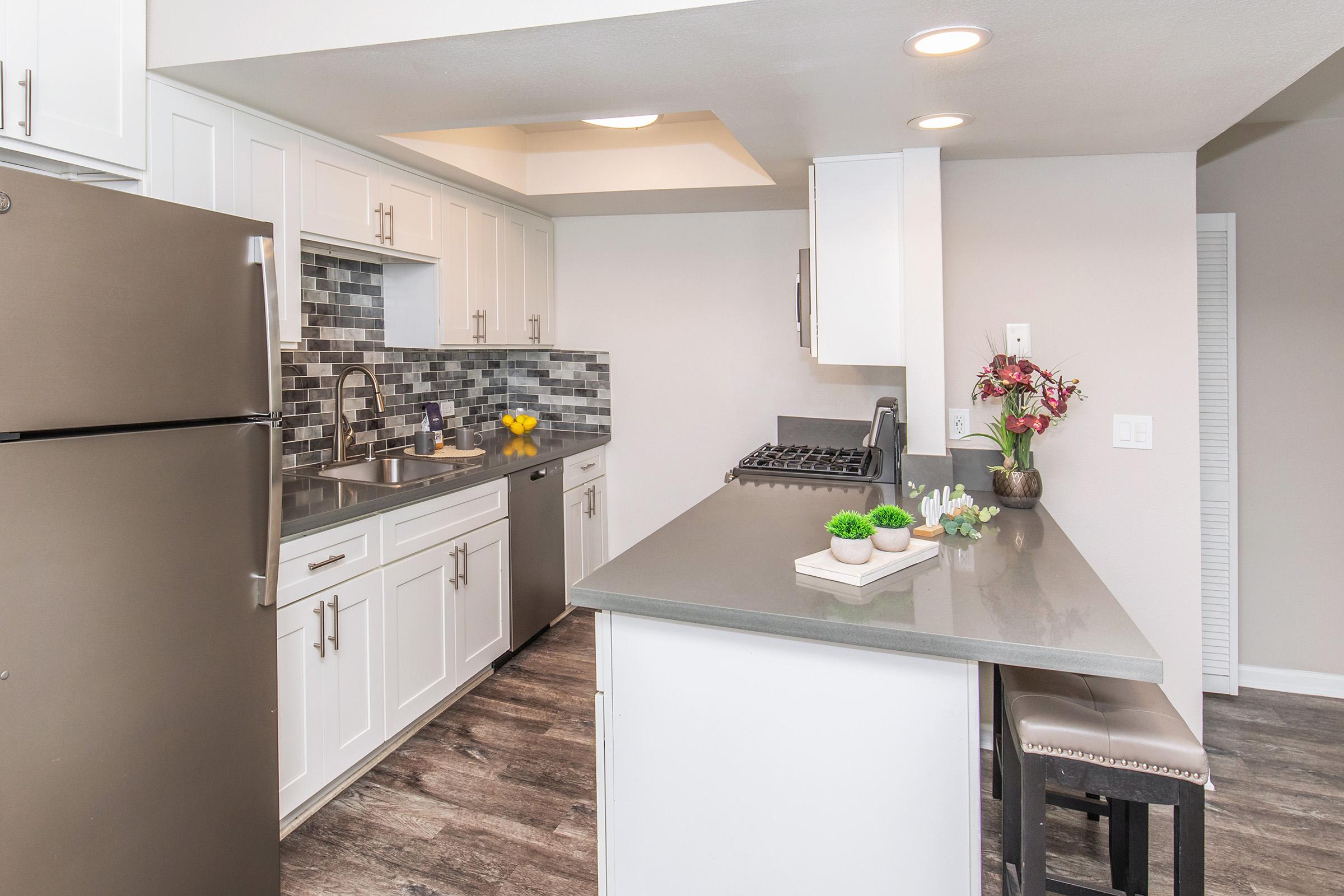 Modern kitchen featuring white cabinetry, stainless steel appliances, and a gray countertop. The backsplash is a mix of gray tiles. A large island with seating is in the center, decorated with small plants and flowers. Natural light brightens the space, enhancing the sleek design and open layout.