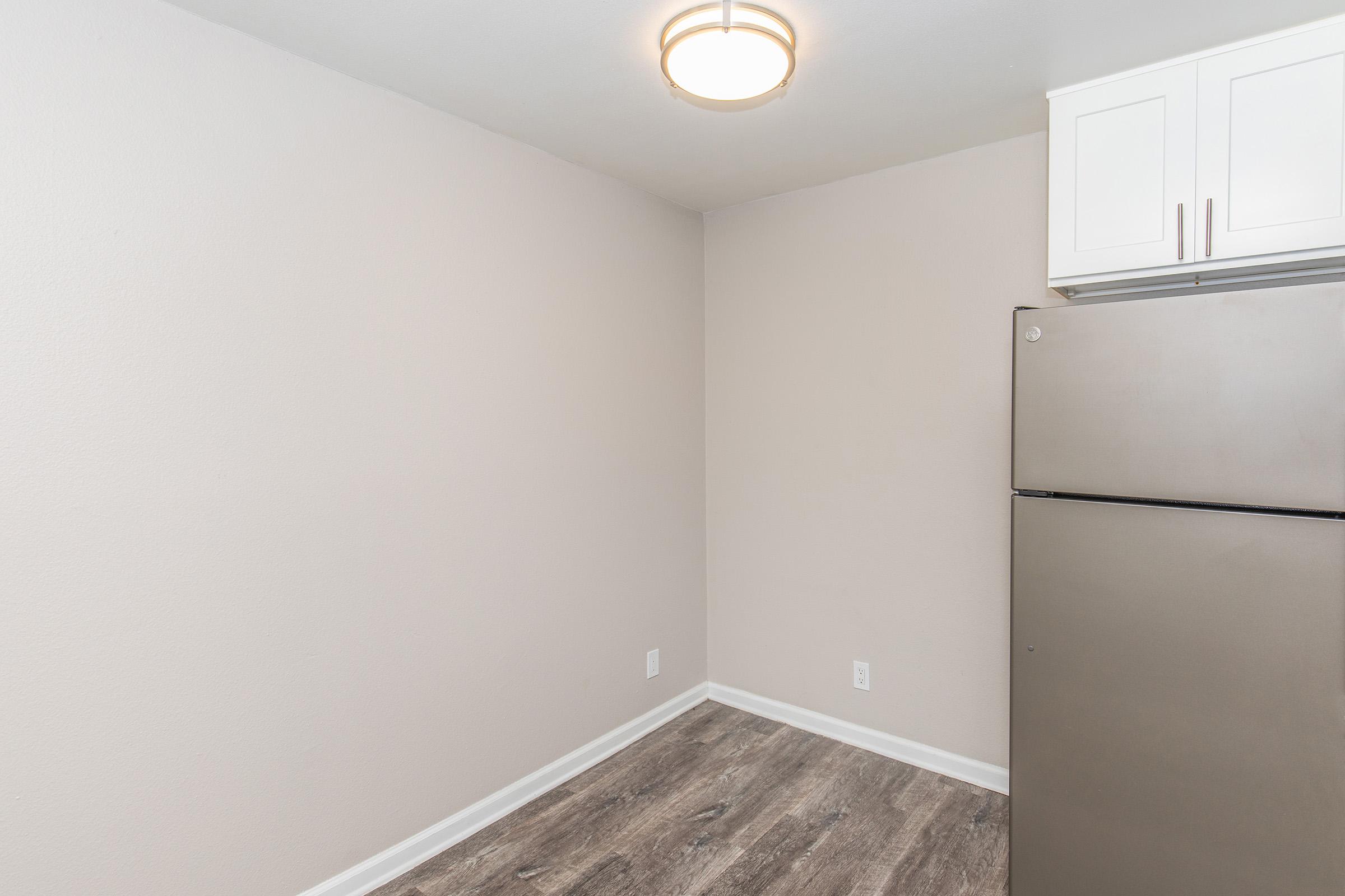 Empty kitchen space featuring a beige wall, a modern ceiling light fixture, a stainless steel refrigerator, and light-colored flooring. The room has minimal furnishings and a clean, simple design, making it suitable for various kitchen styles.