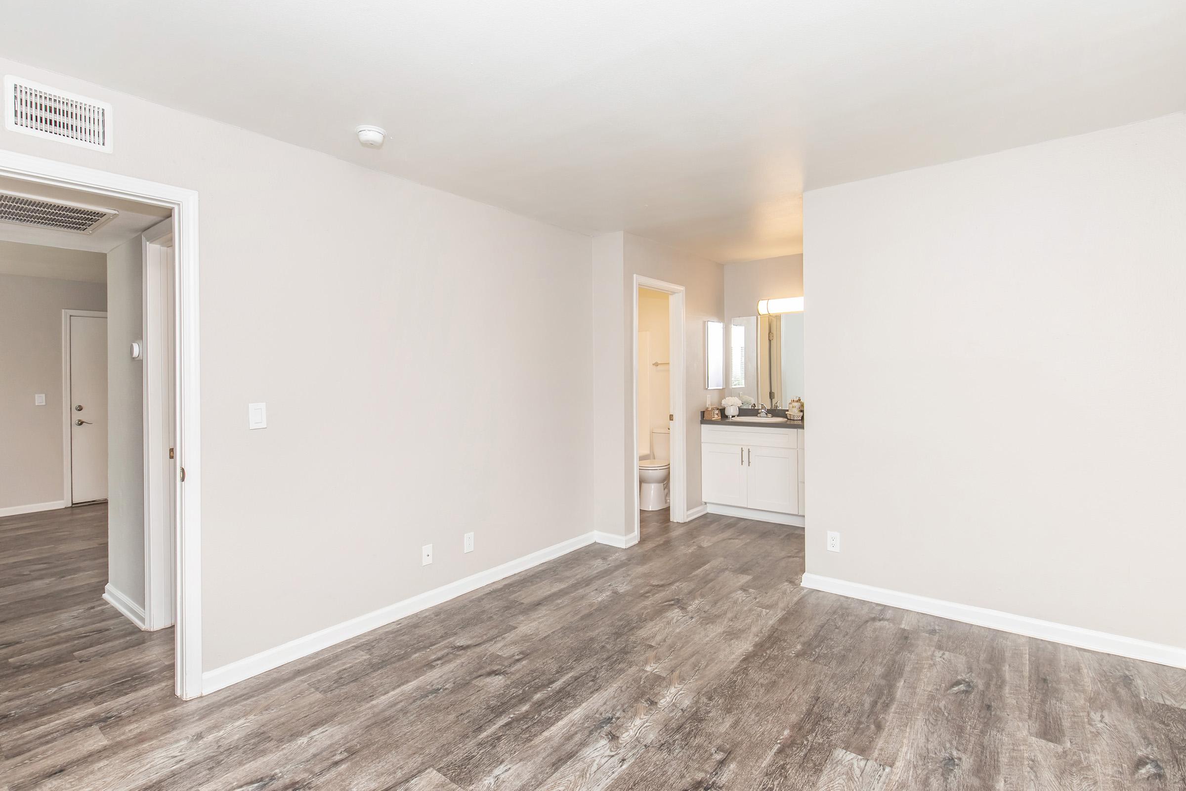 A view of a spacious, empty room with light-colored walls and a modern laminate floor. A doorway leads into a bathroom, which has a vanity and mirror. Natural light illuminates the space, creating a bright and airy atmosphere, suitable for potential furnishings and decor.