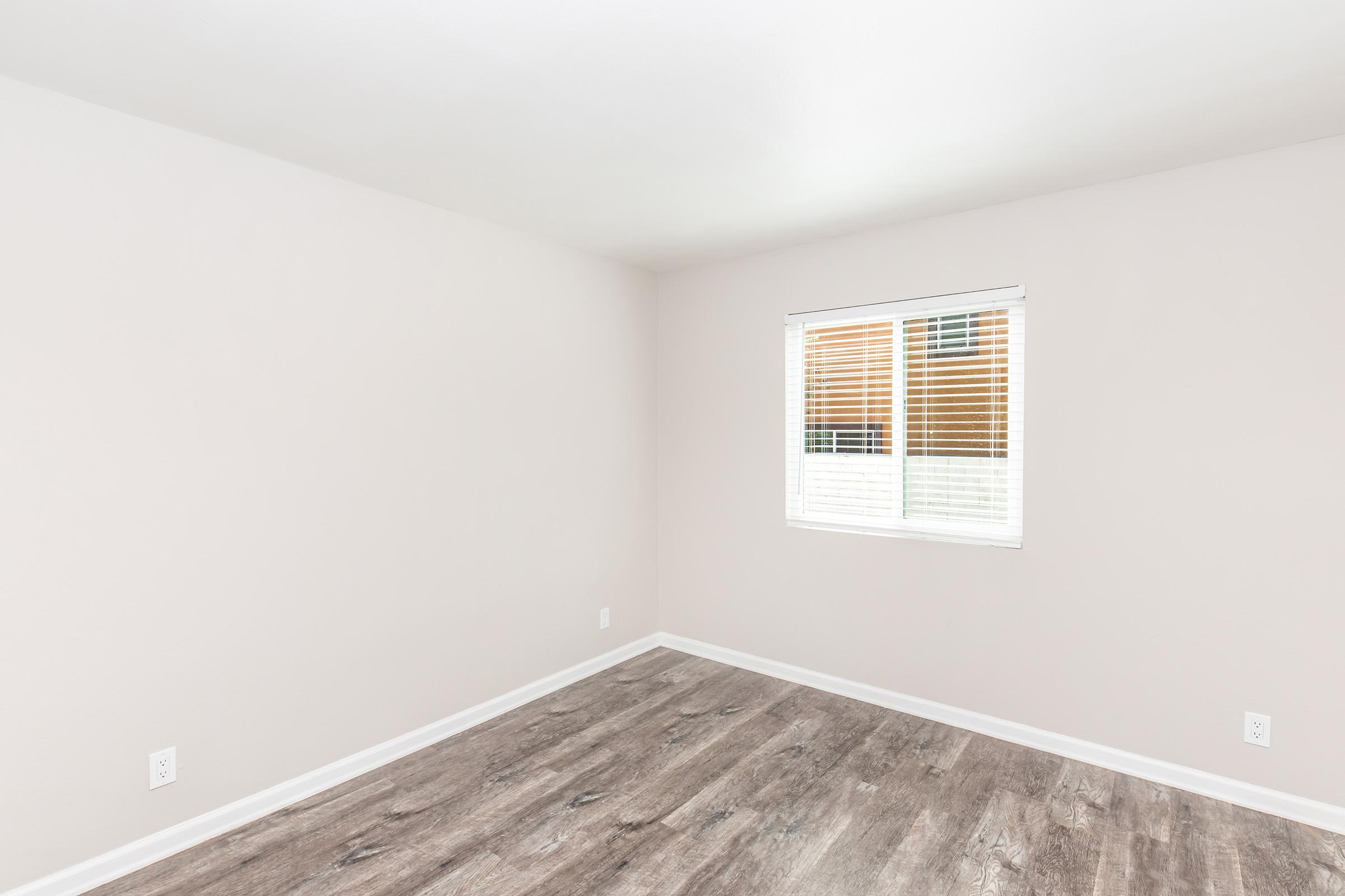 Empty room with light beige walls and a large window featuring white blinds. The floor is made of light-colored wood laminate. Natural light enters through the window, illuminating the space, which has no furniture or decoration. The overall atmosphere is bright and airy.