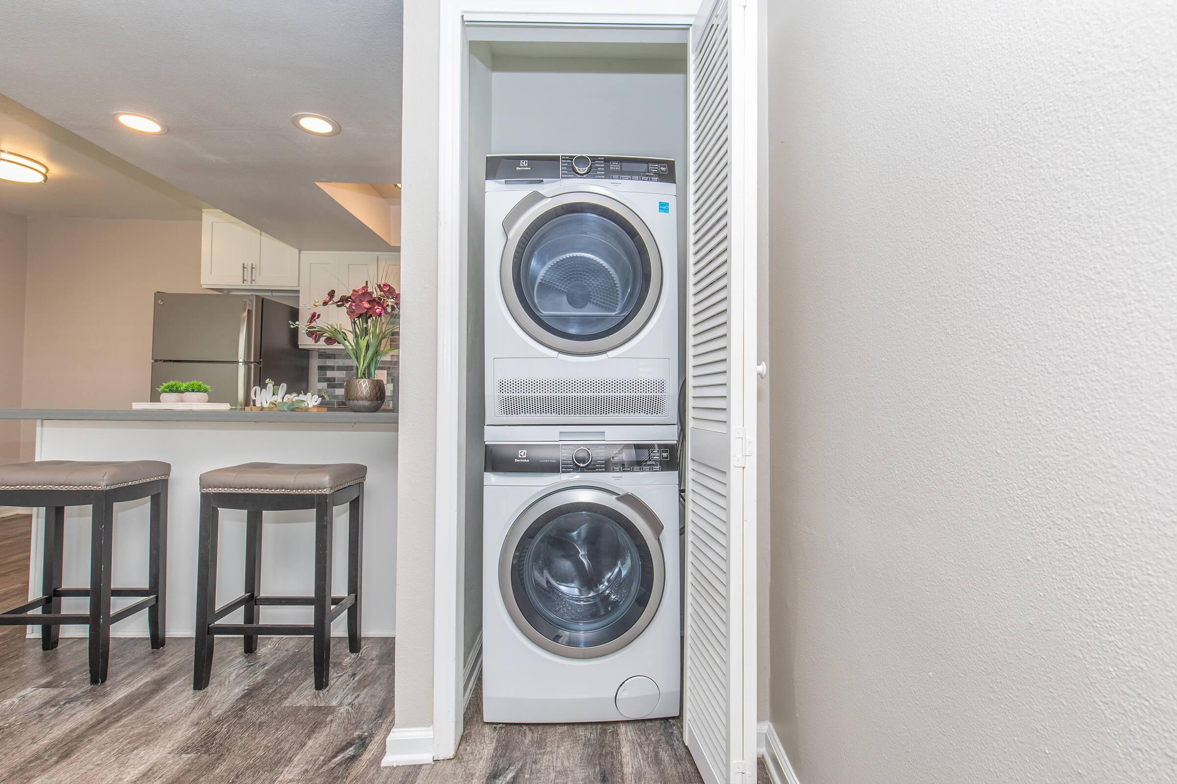 A compact laundry space featuring a stacked washer and dryer in a closet, with modern appliances. The nearby area includes a kitchen with a countertop and two bar stools, and a vase with flowers adds a decorative touch. The setting has neutral-colored walls and a wood-like floor finish.