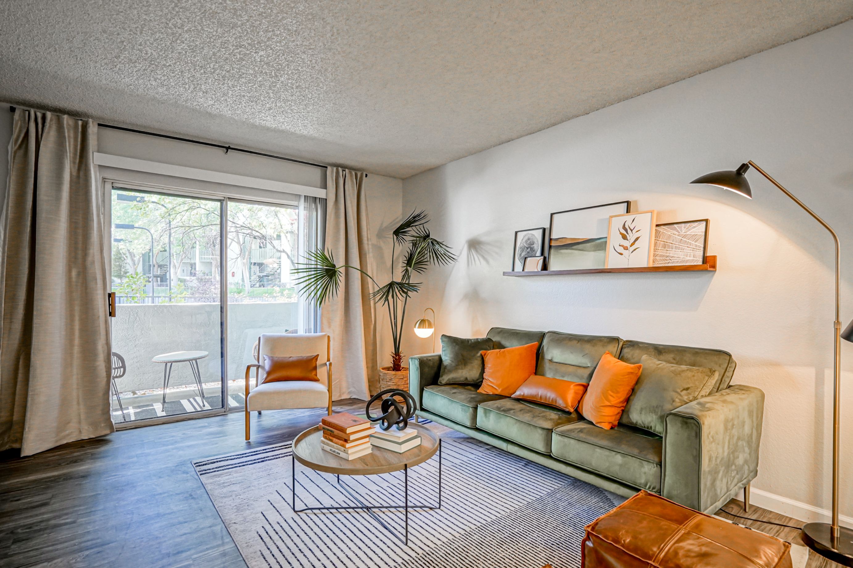 Photograph of a modern living room featuring a green sofa with orange and green cushions, a round coffee table with books and decorative items, and a wooden chair near sliding glass doors leading to a balcony. The room includes a floor lamp, a potted plant, a wall shelf with framed art, and neutral-colored curtains, creating a cozy and stylish atmosphere.