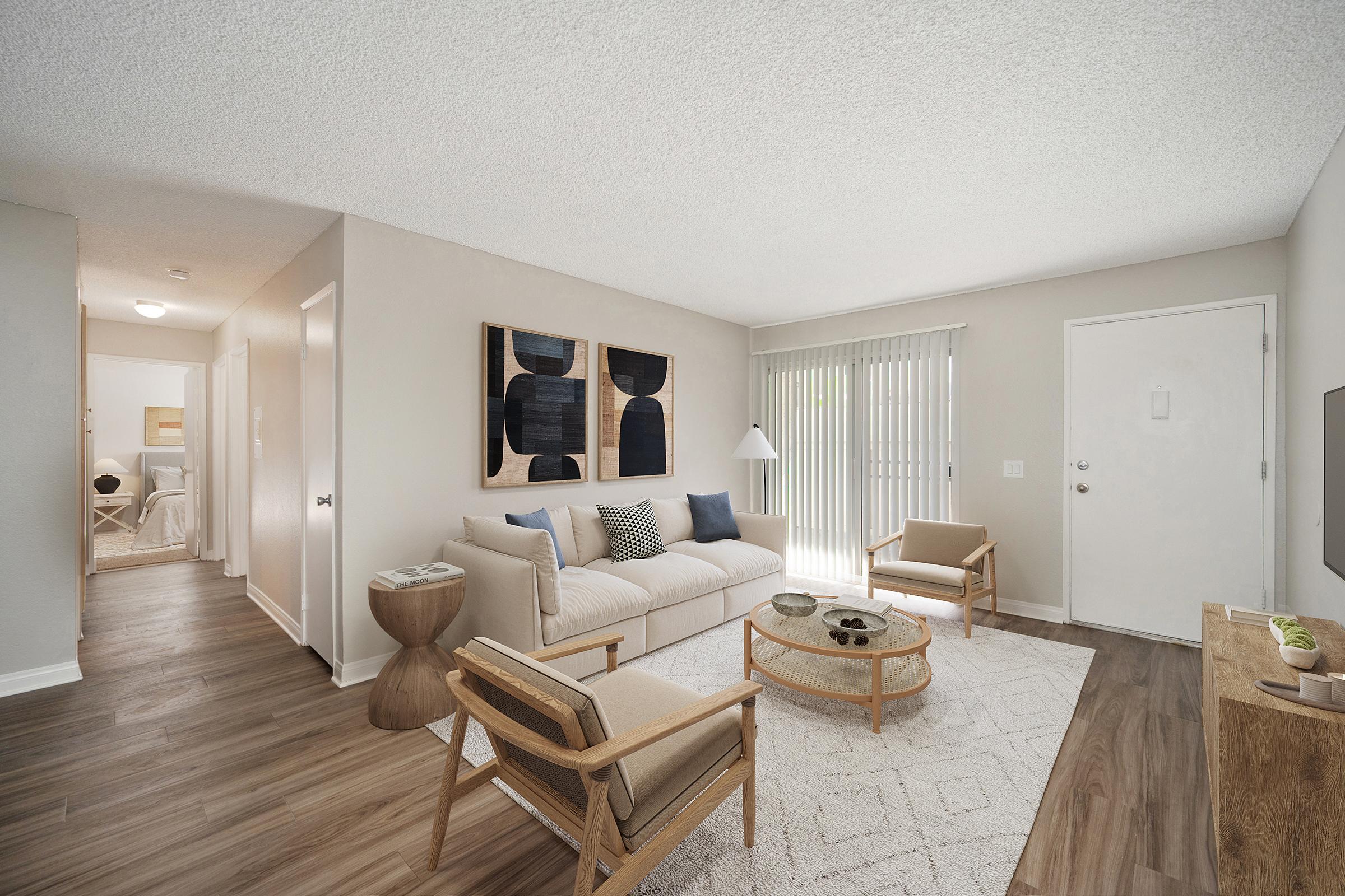 An apartment living room with a beige sofa, two wooden armchairs with tan cushions, and a round woven coffee table on a white patterned rug. Two pieces of abstract black-and-white art hang on light gray walls, and a sliding glass door with vertical blinds lets in natural light.