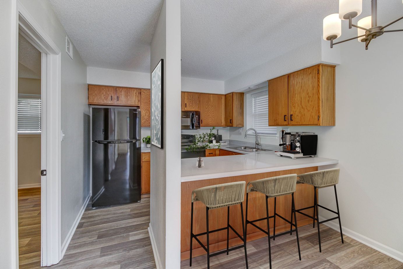 Photograph of a modern kitchen featuring wooden cabinets, a black refrigerator, and a white countertop with three woven bar stools. The space includes a coffee machine, a sink under a window, and light-colored flooring, highlighting a clean and functional design.