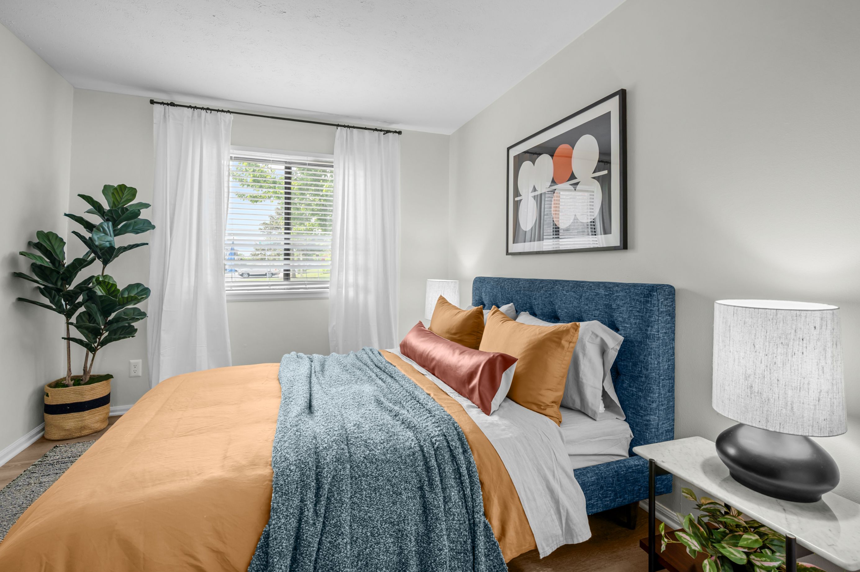 Photograph of a modern bedroom showcasing a neatly made bed with a blue upholstered headboard, mustard yellow and rust-colored pillows, and a blue textured throw blanket. The room features white walls, a large window with white curtains, a potted plant in a woven basket, a bedside table with a lamp, and abstract wall art above the bed.