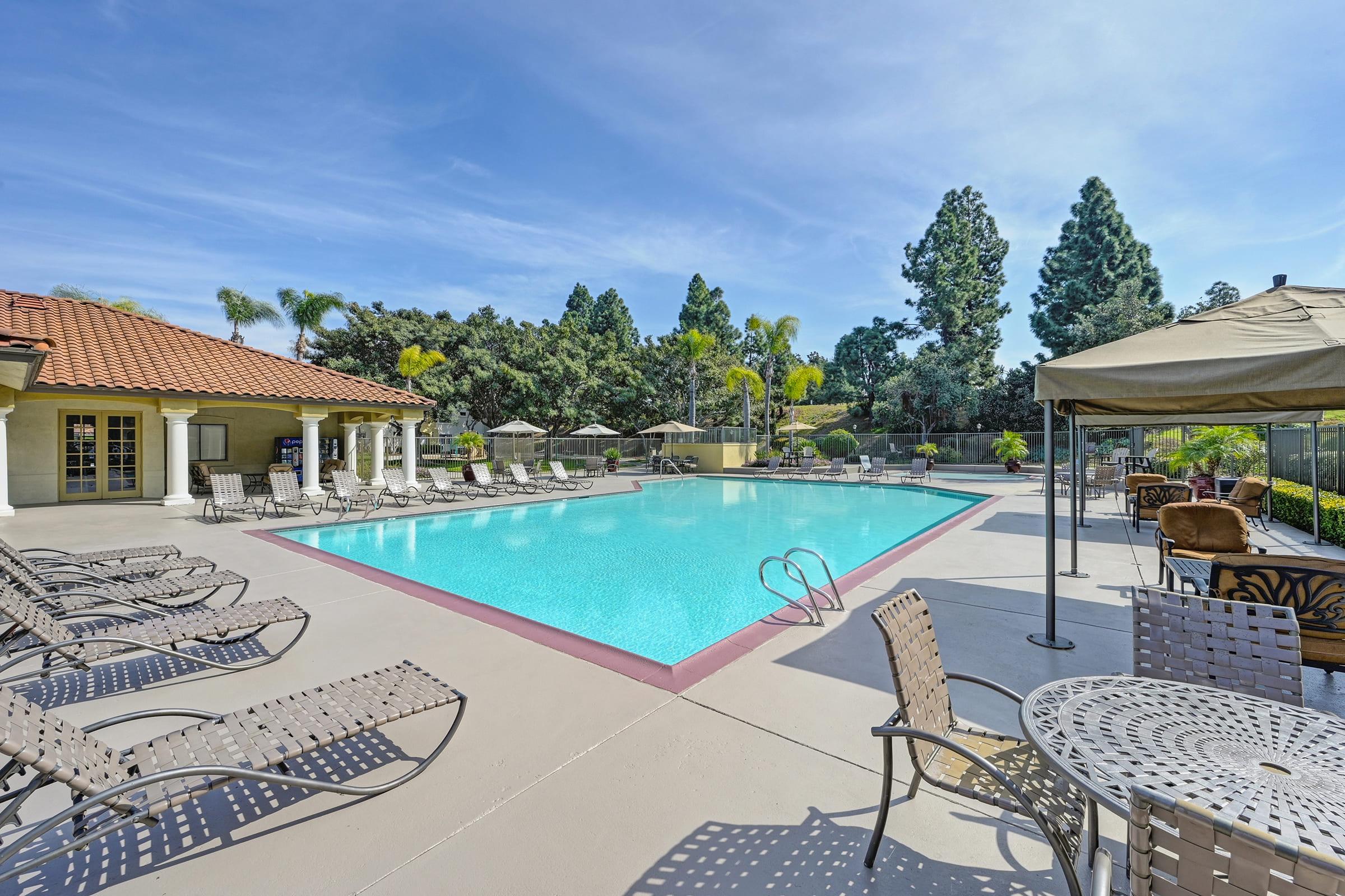 a large pool surrounded by reclining chairs, umbrellas, and tables, in front of a building and green tree-lined background under the blue sky