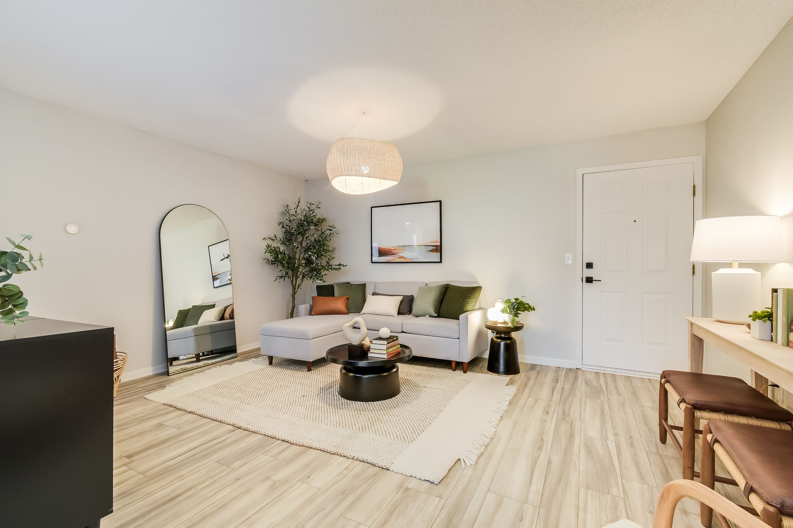 Photograph of a modern living room featuring a light gray sectional sofa with green and brown cushions, a round black coffee table on a textured beige rug, and a large floor mirror leaning against a white wall. The room includes light wood flooring, a white front door, a hanging woven light fixture, a potted plant, and a console table with two brown stools and a lamp.