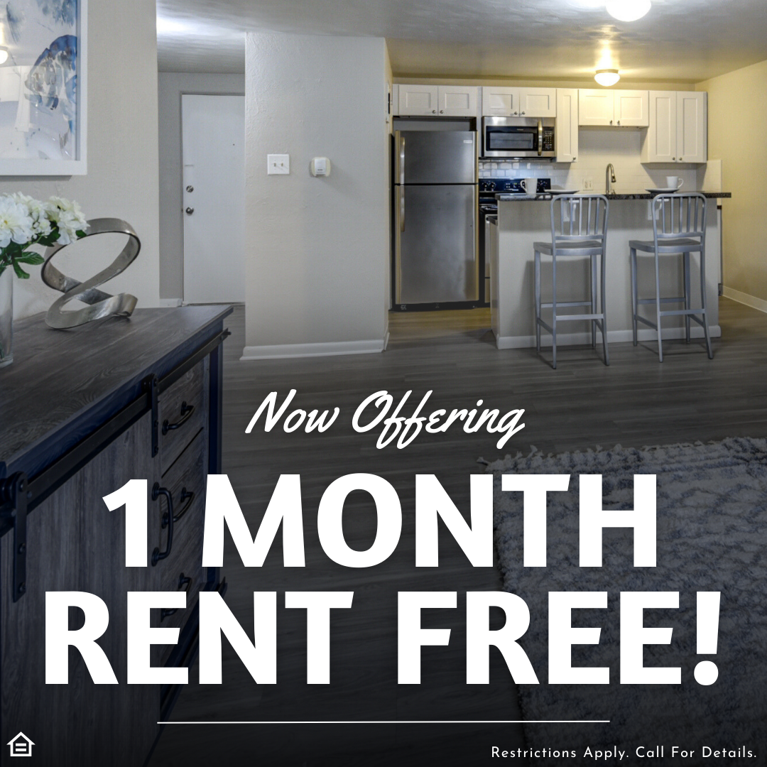Modern apartment interior with open layout featuring a kitchen with stainless steel appliances, white cabinets, bar seating, and a cozy living area. Promotional text over the image reads: “Now Offering 1 Month Rent Free! Restrictions Apply. Call For Details.”