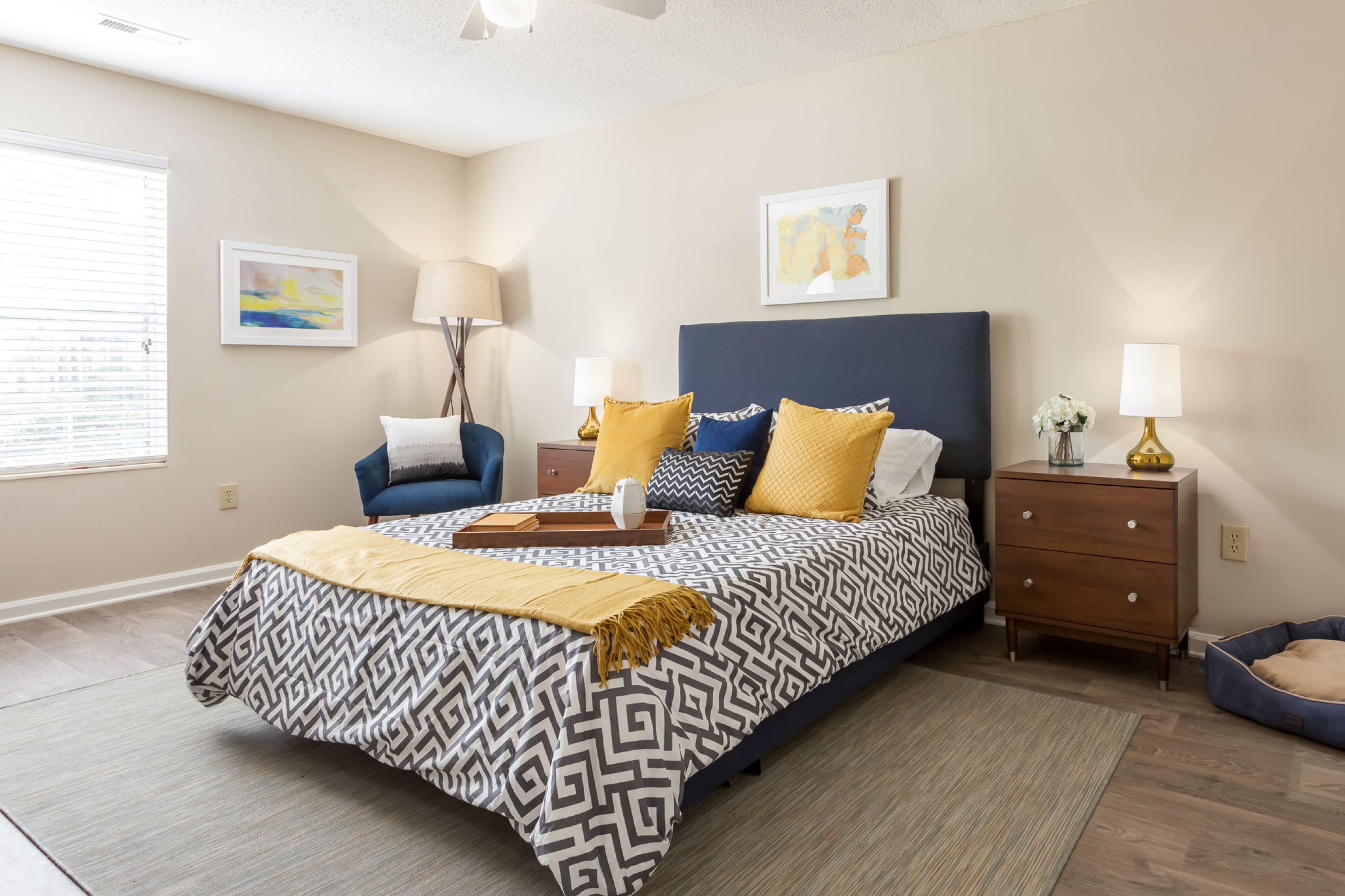 Photograph of a modern bedroom featuring a queen-sized bed with a navy blue headboard, geometric patterned bedding, and yellow accent pillows. Room includes a wooden nightstand with a lamp and flowers, a blue armchair with a white pillow, framed artwork on beige walls, and a window with blinds allowing natural light.