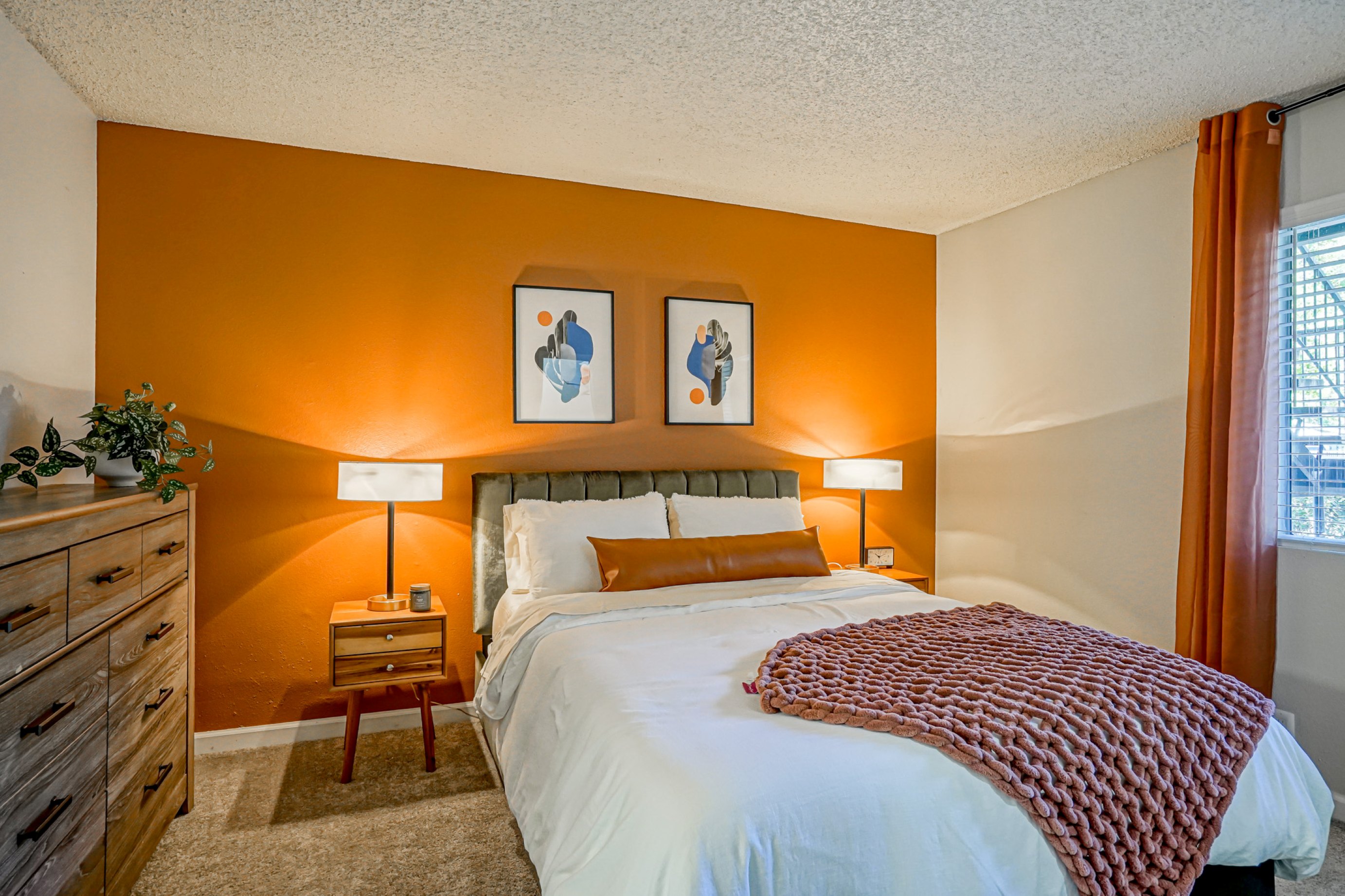 Photograph of a modern bedroom featuring a bed with white linens, a rust-colored throw blanket, and a matching long pillow against an orange accent wall. Room includes two wooden nightstands with lamps, a wooden dresser with a plant, abstract artwork above the bed, and a window with orange curtains allowing natural light.