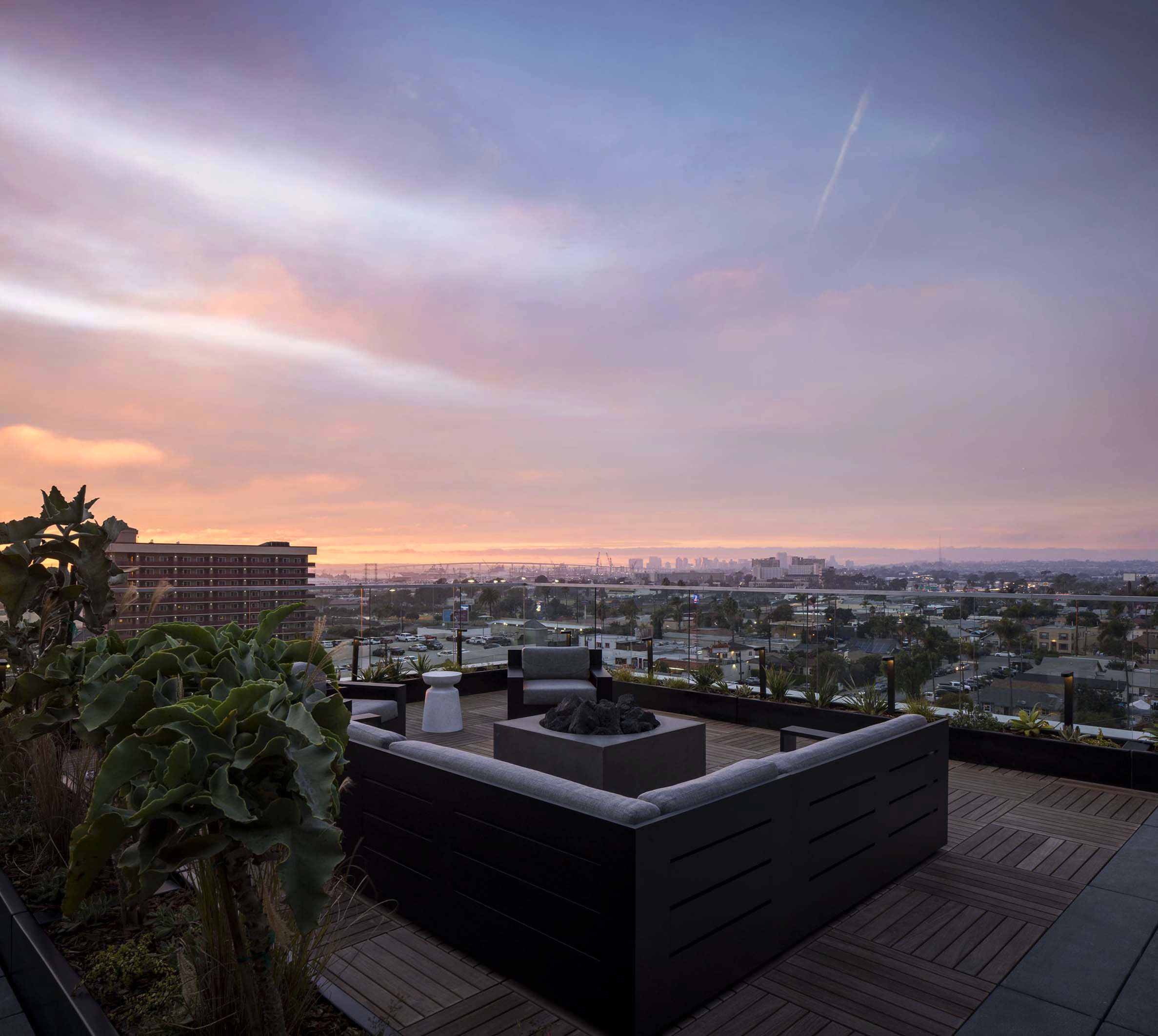 a rooftop lounge with a couch and 2 chairs circling a fire pit on wood plank flooring, next to green plants in front of a sunset skyline view of downtown San Diego