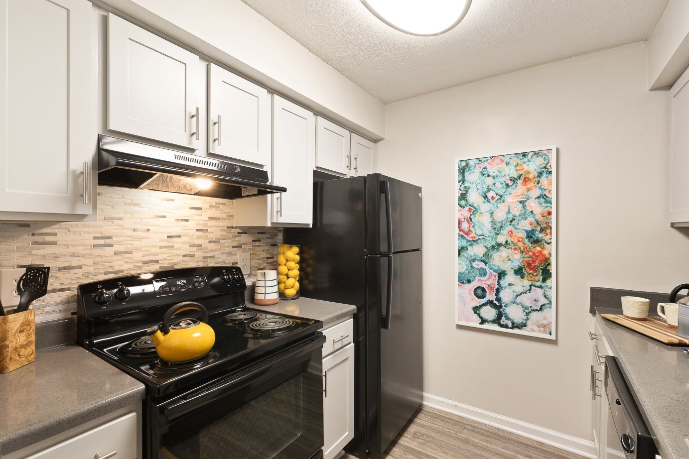 Photograph of a modern kitchen featuring white cabinets, a black refrigerator, and a black stove with a yellow kettle on top. The space includes a beige tiled backsplash, gray countertops, and a colorful abstract painting on a white wall.